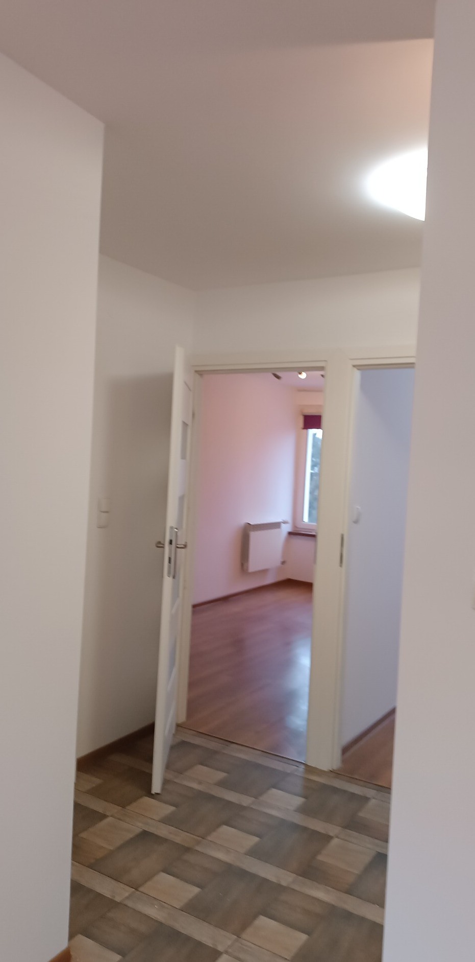 Widok na odnowione wnętrze mieszkania z korytarza: podłoga wyłożona panelami imitującymi drewno, białe ściany i drzwi, w tle pokój z drewnianą podłogą i grzejnikiem pod oknem.