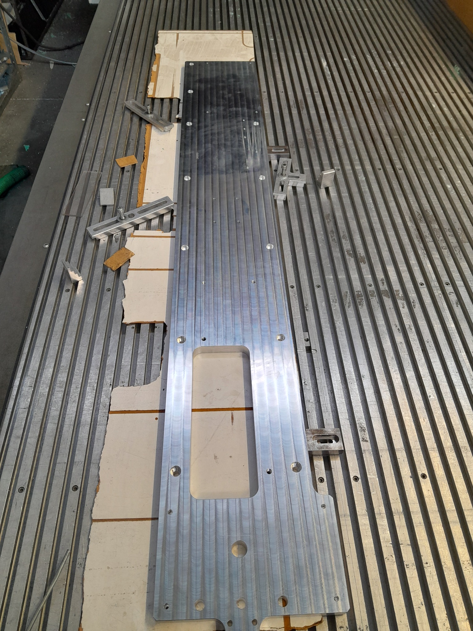 Detal z obrobionego metalu, przymocowany do stołu z rowkami teowymi za pomocą uchwytów, widoczne resztki materiału pod spodem.