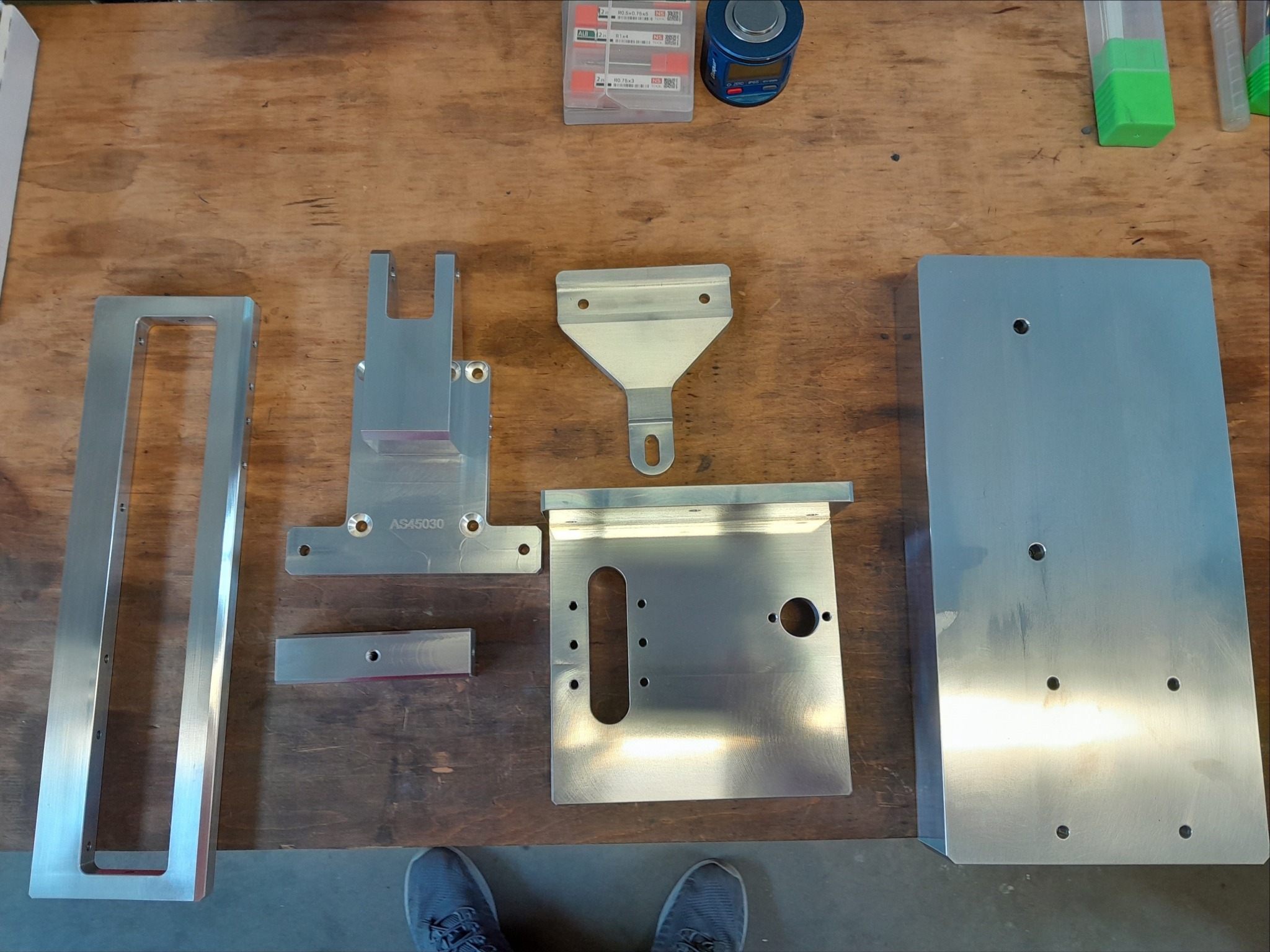 Precyzyjnie obrobione, srebrne, metalowe elementy o różnych kształtach i rozmiarach leżące na drewnianym stole warsztatowym, wraz z narzędziami i pudełkami w tle.