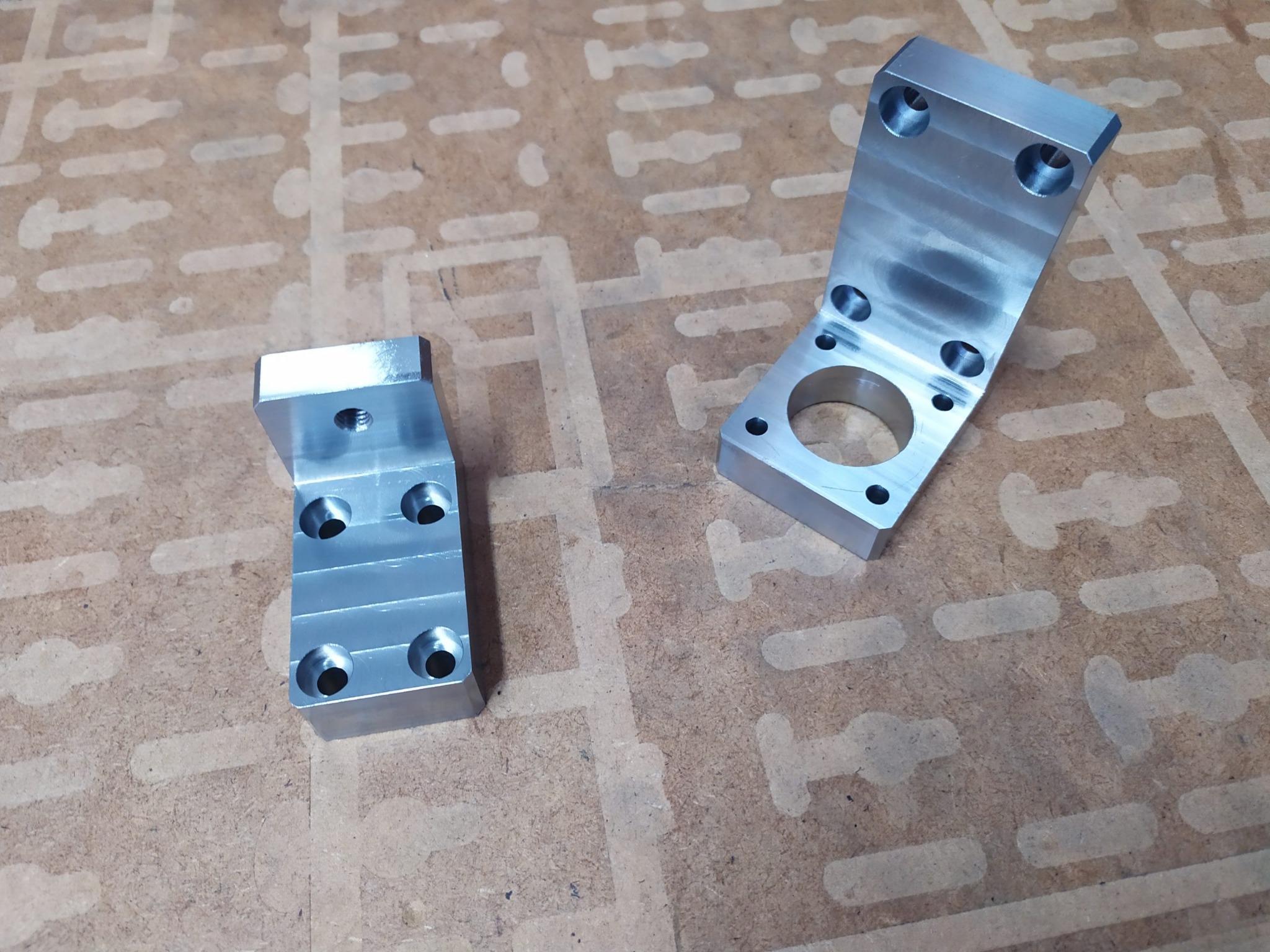 Dwie metalowe, obrobione maszynowo, srebrne kątowniki z otworami, leżące na brązowym blacie z wzorem. Większy kątownik ma otwór centralny.