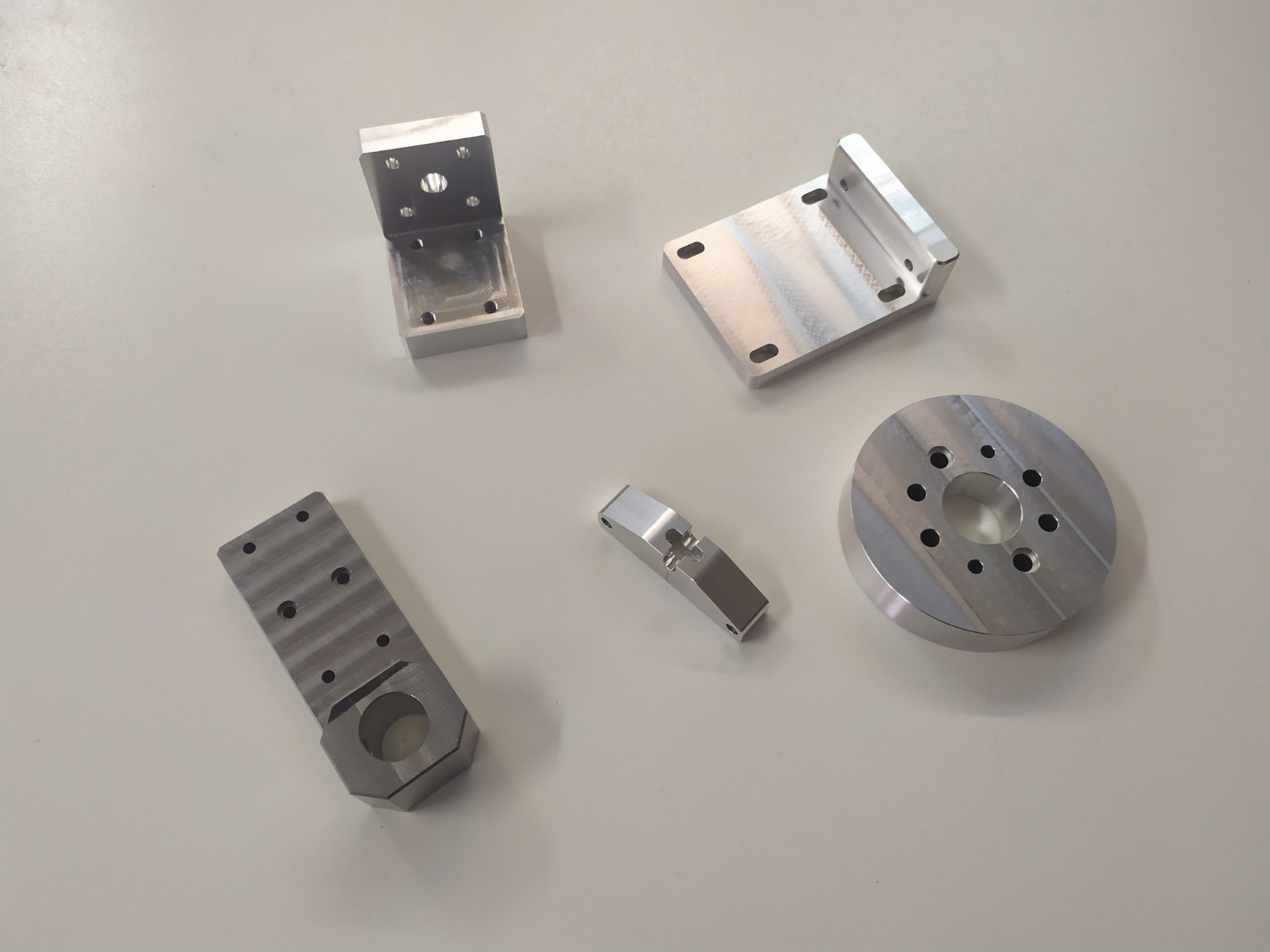 Precyzyjnie obrobione, srebrne, metalowe elementy o różnych kształtach, w tym kątownik, płyta z otworami i cylinder, ułożone na gładkim, jasnym tle.