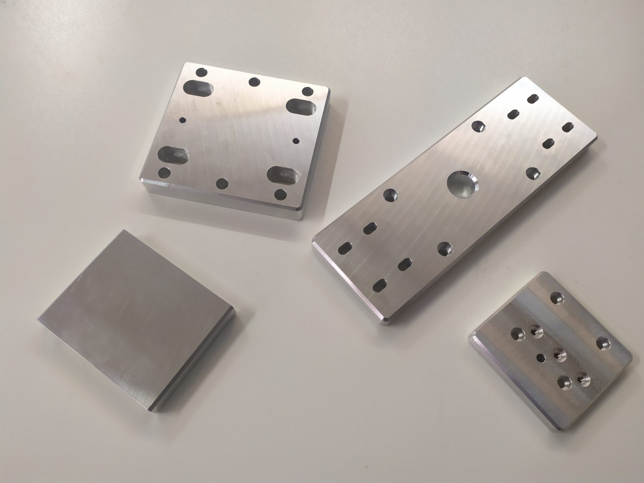 Cztery precyzyjnie obrobione, srebrne elementy metalowe o różnych kształtach i rozmiarach, z licznymi otworami i frezowaniami, ułożone na jasnym, gładkim tle. Widoczne ślady obróbki skrawaniem.