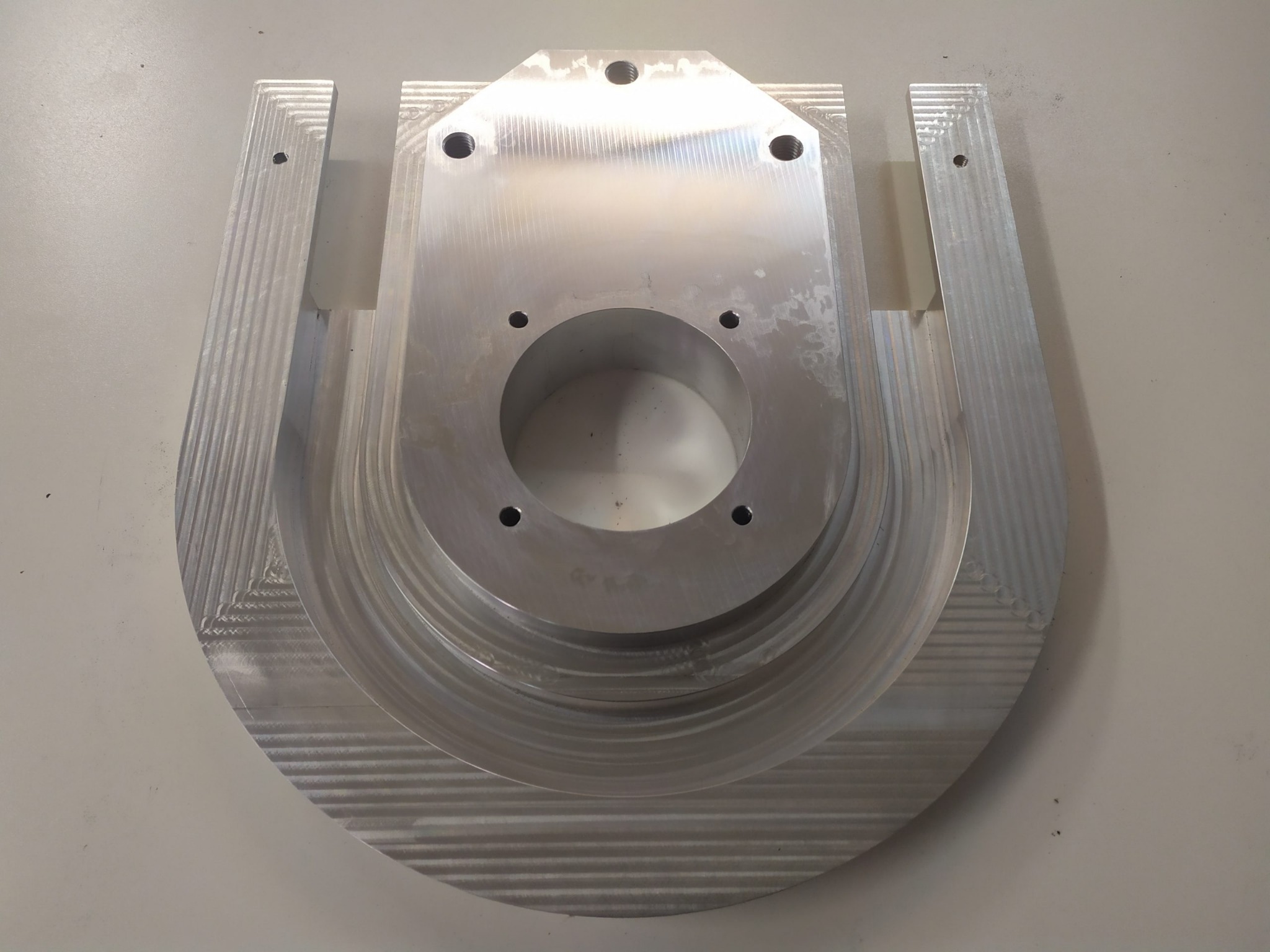 Metalowy element obrobiony z widocznymi śladami frezowania, otwory montażowe i kształt przypominający podkowę z centralnym otworem.