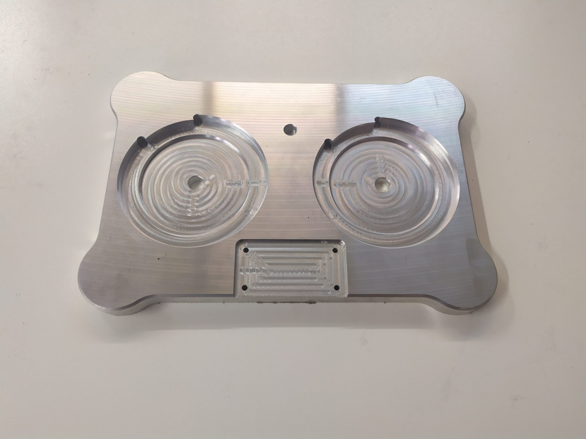 Aluminiowy element po obróbce CNC z dwoma okrągłymi wgłębieniami z koncentrycznymi rowkami i prostokątnym wyżłobieniem z meandrycznym wzorem, leżący na jasnym tle.