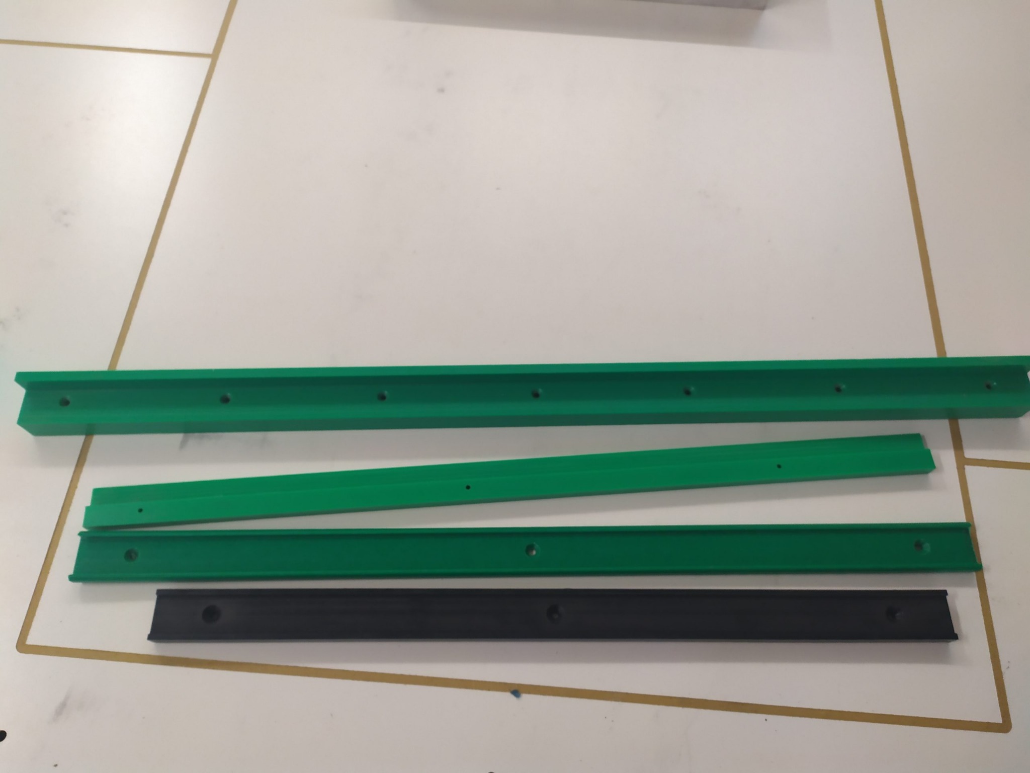 Zielone i czarne, profilowane listwy z otworami, ułożone równolegle na białym stole z brązowymi obramowaniami.