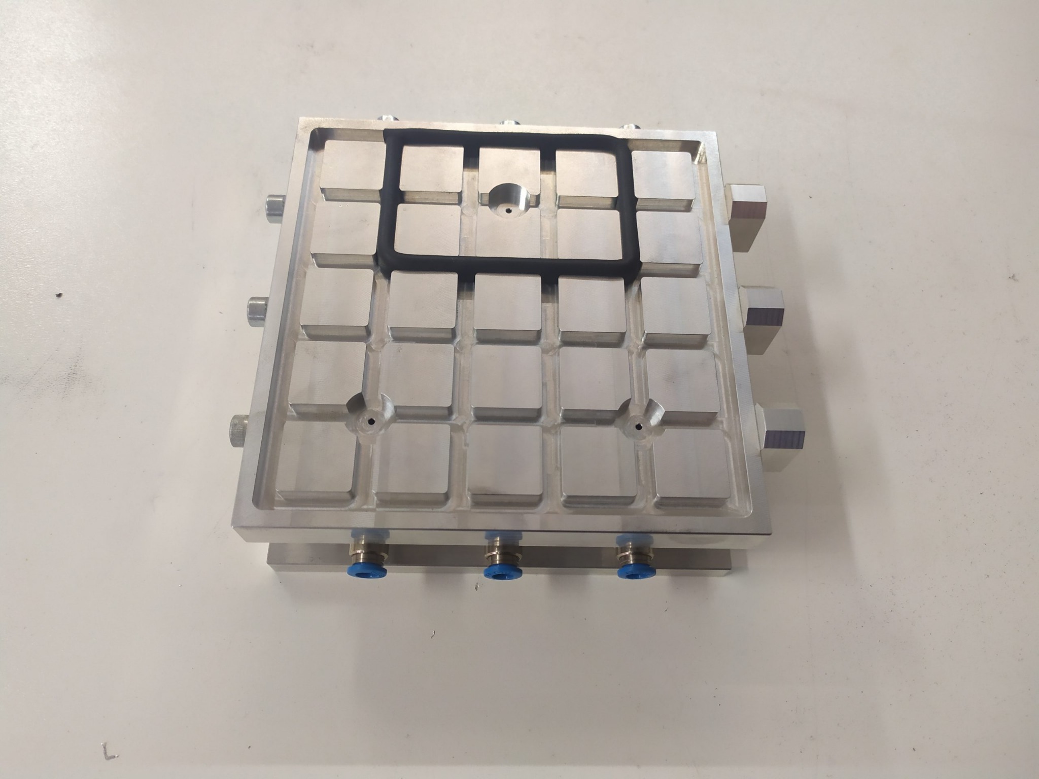 Precyzyjnie obrobiony, srebrny element metalowy z licznymi wgłębieniami w kształcie kwadratów i okrągłymi otworami, prawdopodobnie element maszyny, z widocznymi przyłączami pneumatycznymi...