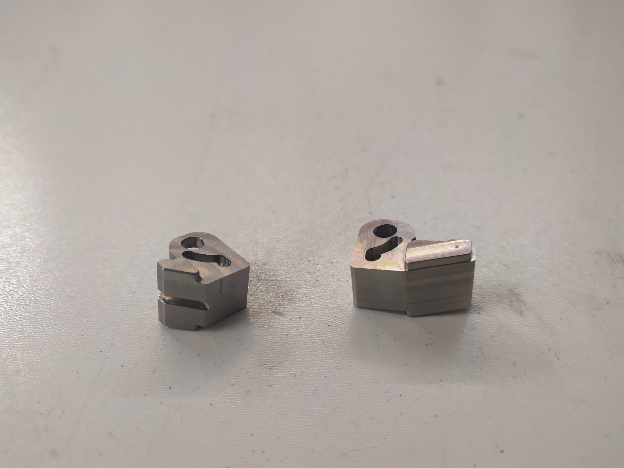 Dwa metalowe elementy z wywierconymi otworami i frezowanymi krawędziami leżą na szarym stole. Jeden element stoi pionowo, a drugi leży na boku, ukazując warstwową strukturę.