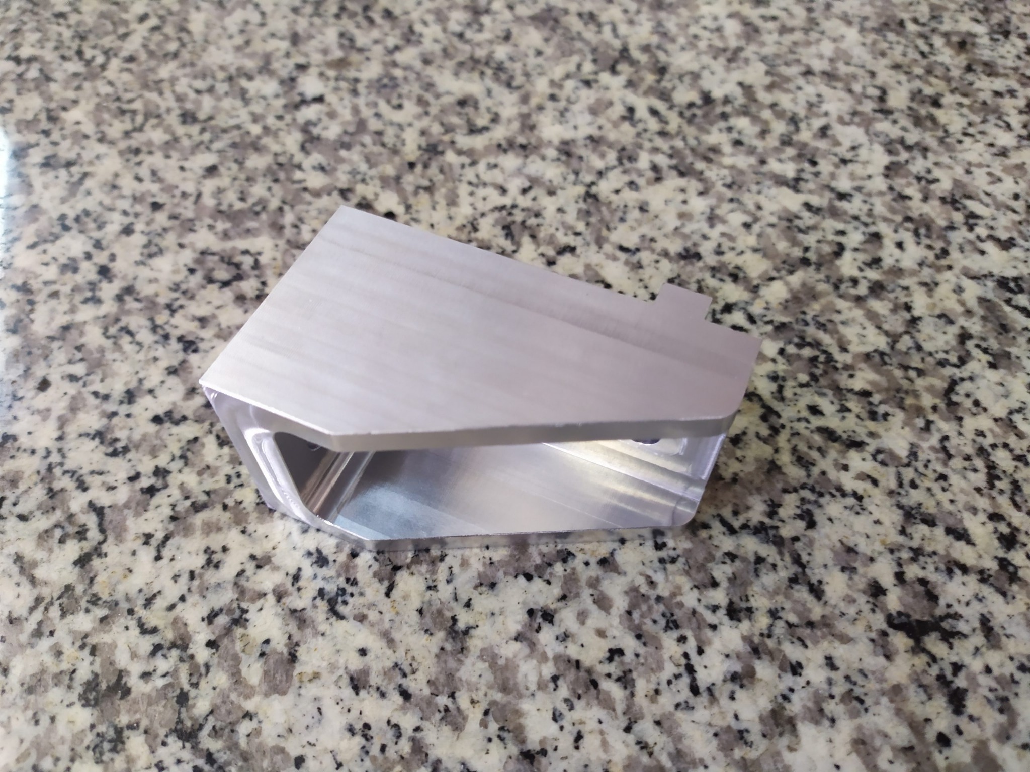 Precyzyjnie obrobiony, aluminiowy element o złożonym kształcie, leżący na blacie z szarego, nakrapianego kamienia. Widoczne ślady obróbki skrawaniem i gładkie powierzchnie.