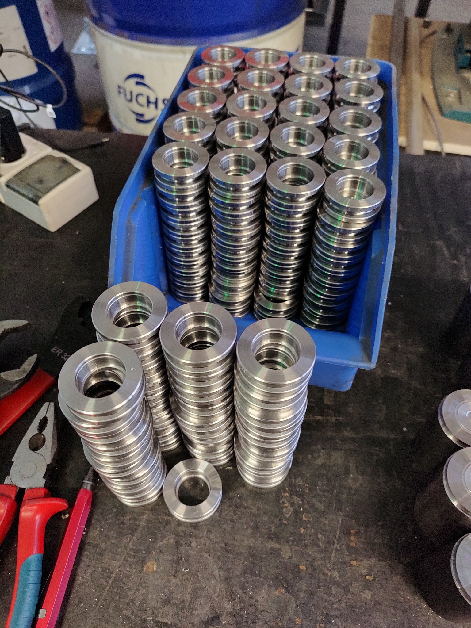 Stosy precyzyjnie obrobionych, metalowych pierścieni w niebieskim pojemniku, ułożone obok narzędzi na warsztatowym stole, z widocznym logo producenta smarów w tle.