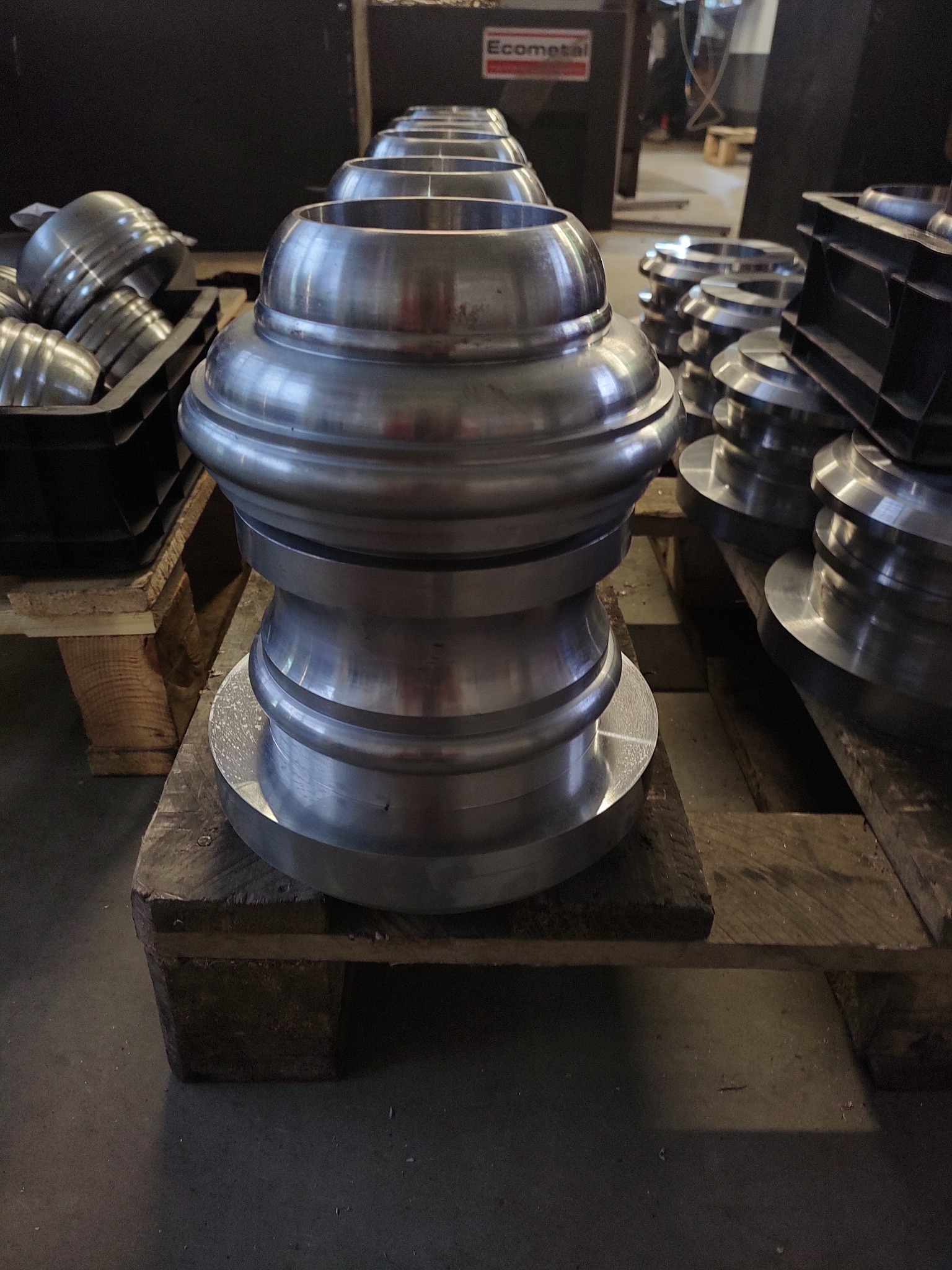 Rząd błyszczących, metalowych elementów o kształcie zbliżonym do klepsydry, umieszczonych na drewnianych paletach w hali produkcyjnej, z widocznym logo 'Ecometal' w tle.