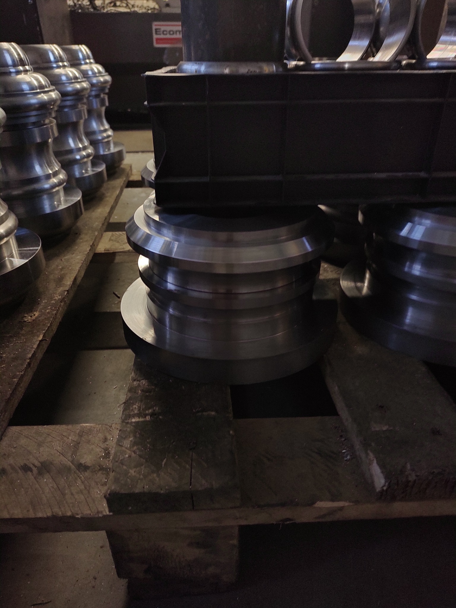 Szereg obrobionych, metalowych elementów o kształcie zbliżonym do kołnierzy, ułożonych na drewnianej palecie. Widoczne ślady obróbki i refleksy światła na powierzchni metalu.