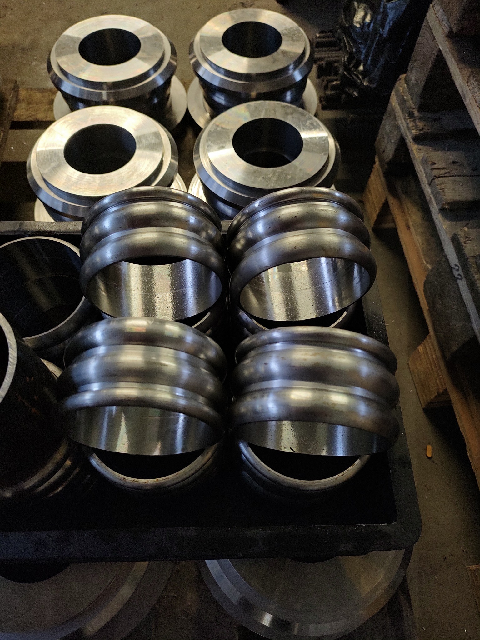 Rzędy metalowych pierścieni i tulei o różnych kształtach i rozmiarach, ułożonych na palecie i w czarnej skrzynce, prawdopodobnie po obróbce.