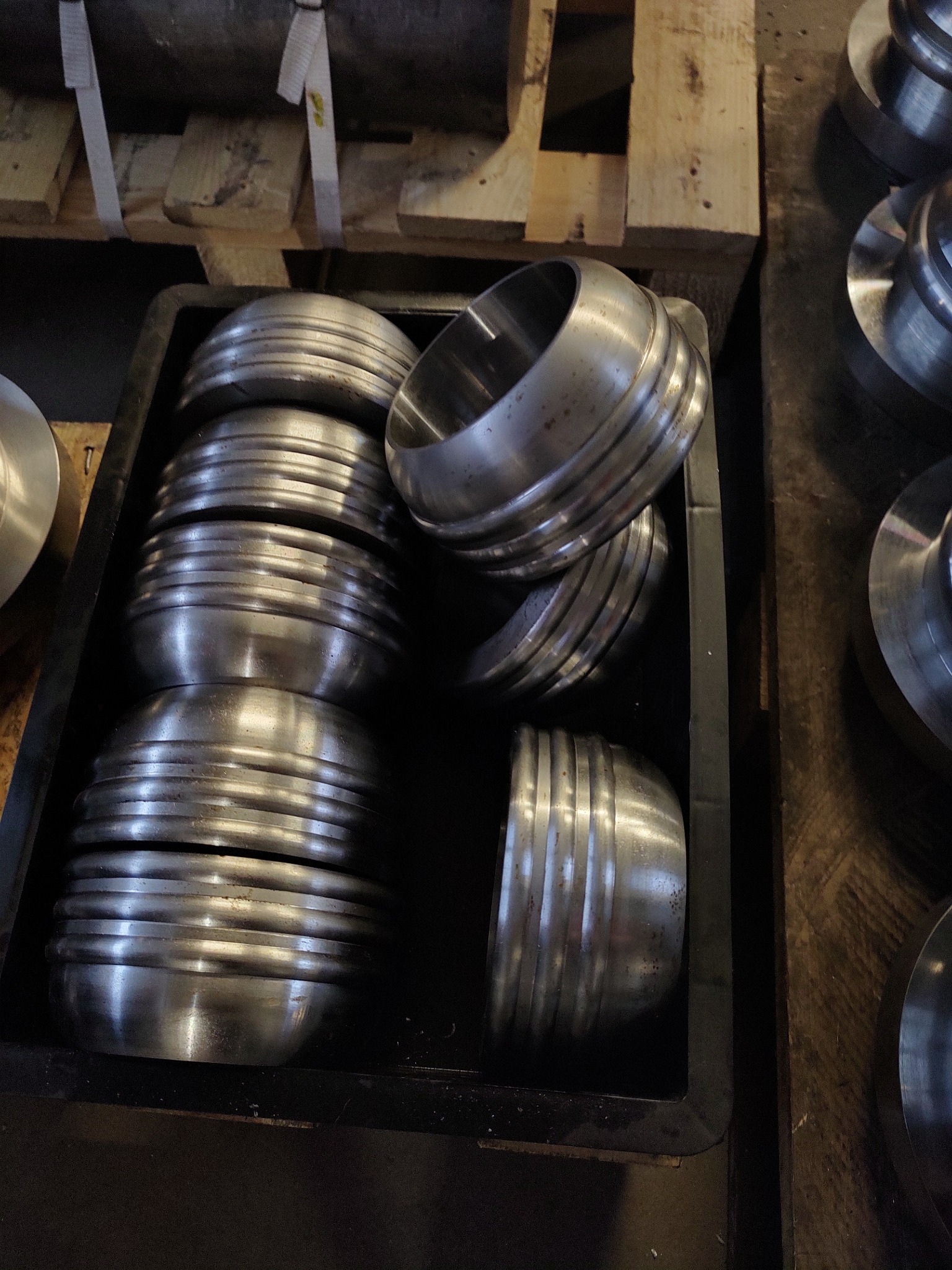 Stos obrobionych, metalowych elementów w kształcie miseczek, ułożonych w czarnej skrzynce transportowej, widoczne ślady obróbki i drobne zarysowania na powierzchni.