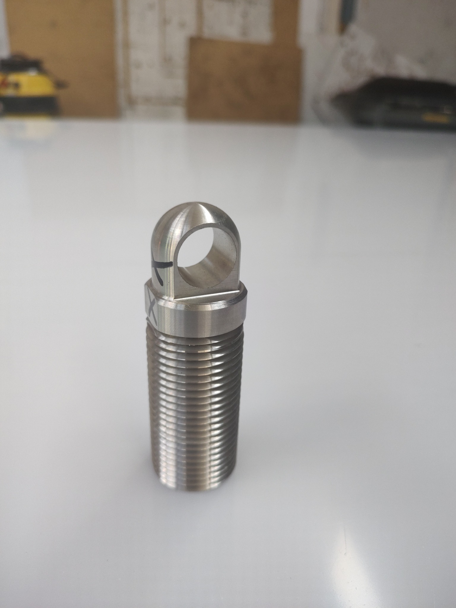 Metalowy element gwintowany z oczkiem, obrobiony na tokarce lub CNC, z widocznymi śladami obróbki i oznaczeniami na powierzchni, leżący na białym blacie.