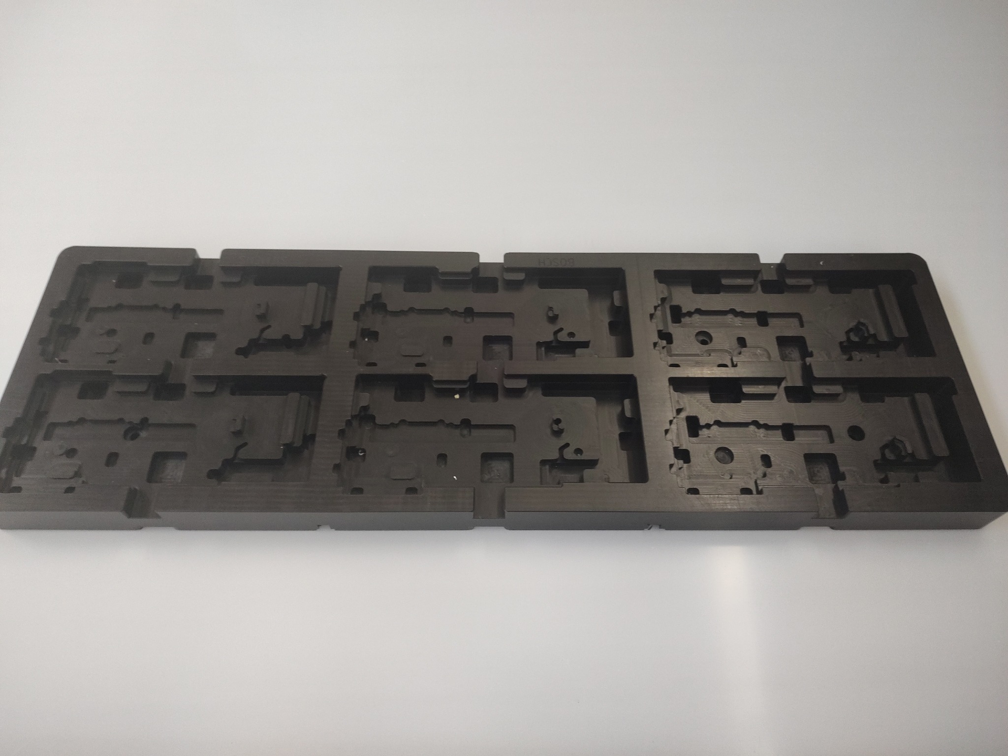 Czarny, precyzyjnie obrobiony element z tworzywa sztucznego z licznymi wgłębieniami i otworami, leżący na białym tle.