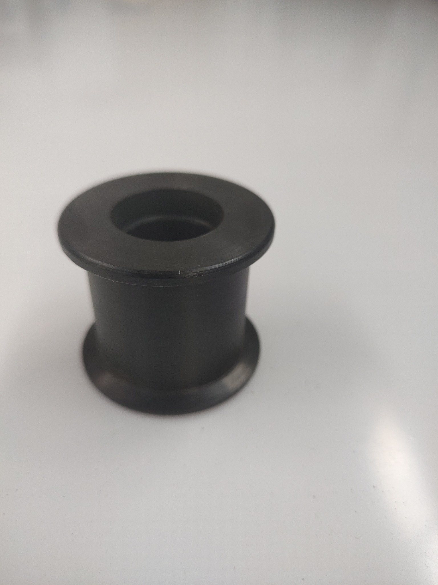 Czarny, metalowy element tulejowy z kołnierzem, wykonany na tokarce lub CNC, leżący na białym tle.
