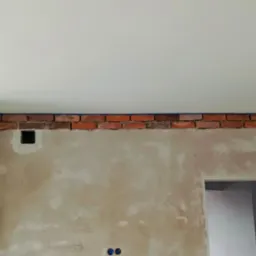 Ściana w trakcie prac wykończeniowych: częściowo położona cegła dekoracyjna obramowana niebieską taśmą malarską, surowy tynk z odręcznymi notatkami i otworem na instalację elektryczną, widoczny...