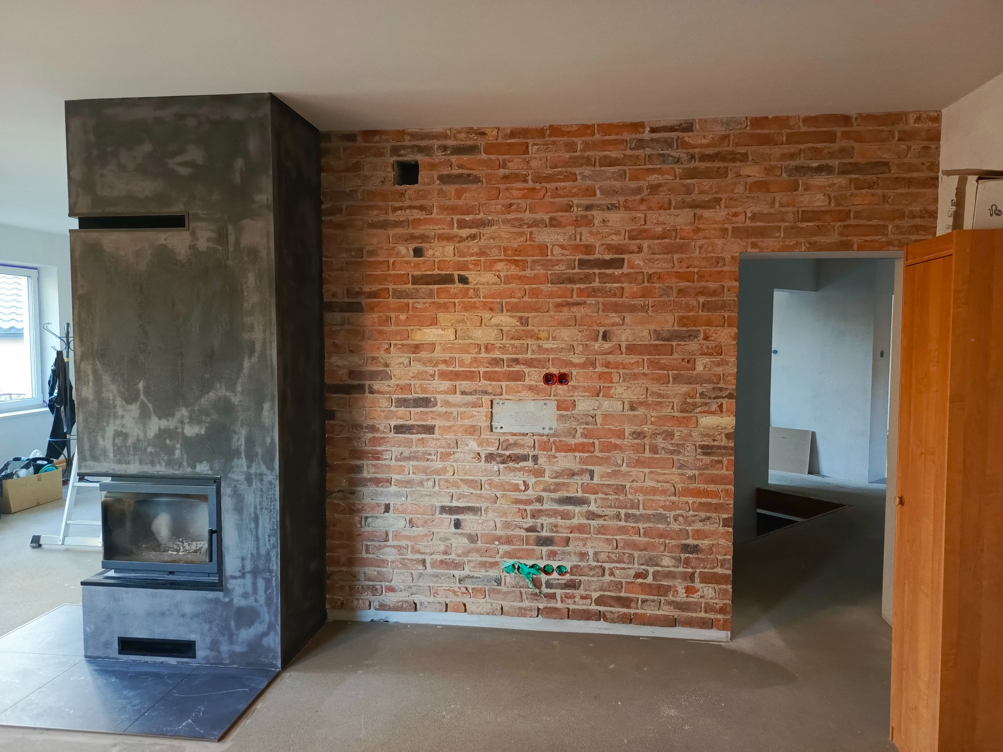Wnętrze w trakcie wykańczania: ściana z czerwonej cegły z widocznymi przewodami, obok ciemny kominek z szarą wylewką, surowa podłoga, widoczny fragment drzwi i przejście do kolejnego pomieszczenia.