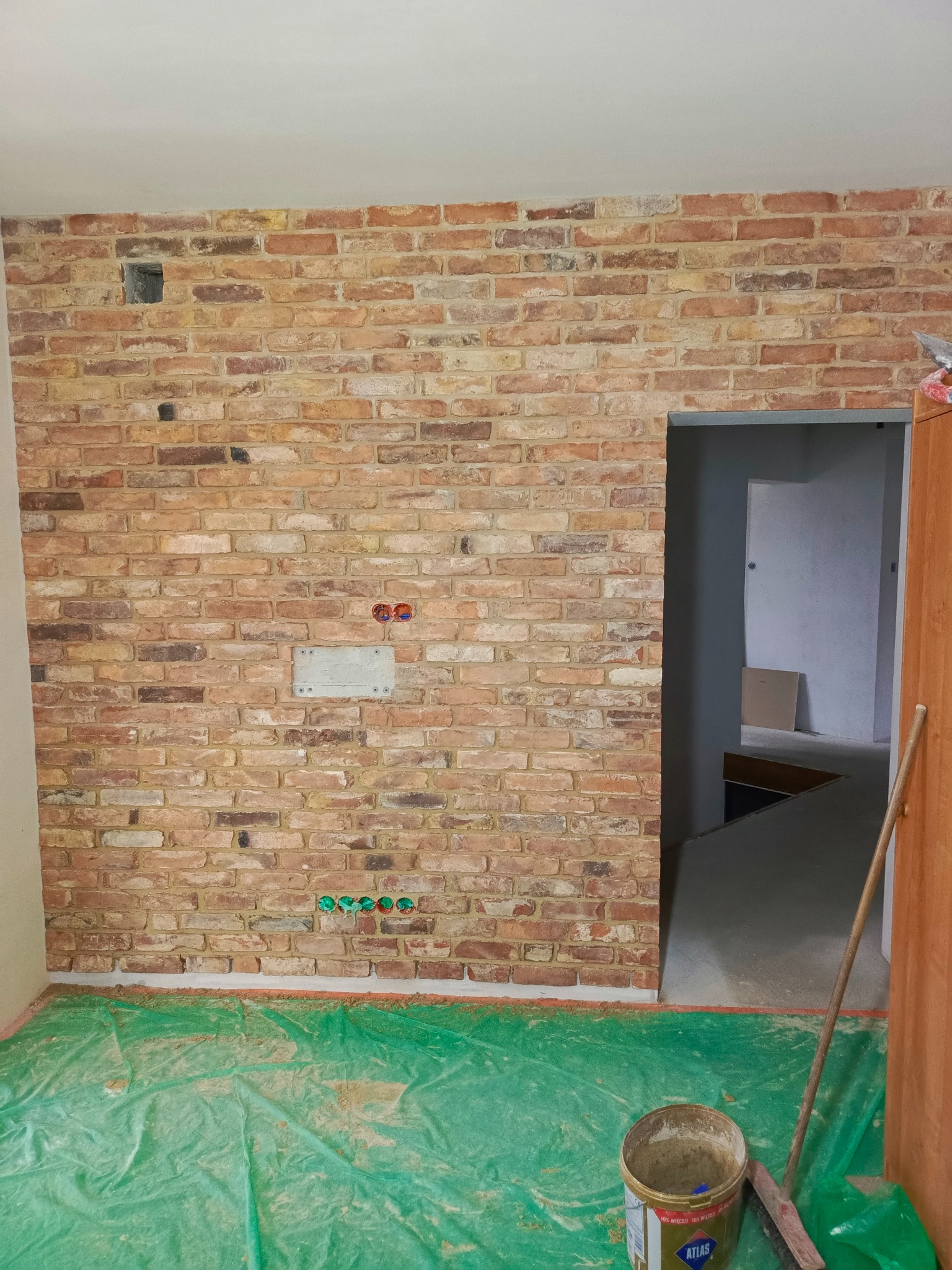 Ściana z cegły klinkierowej w trakcie prac wykończeniowych, widoczne puszki elektryczne, otwór w ścianie, podłoga zabezpieczona folią, wiadro z zaprawą i szczotka.
