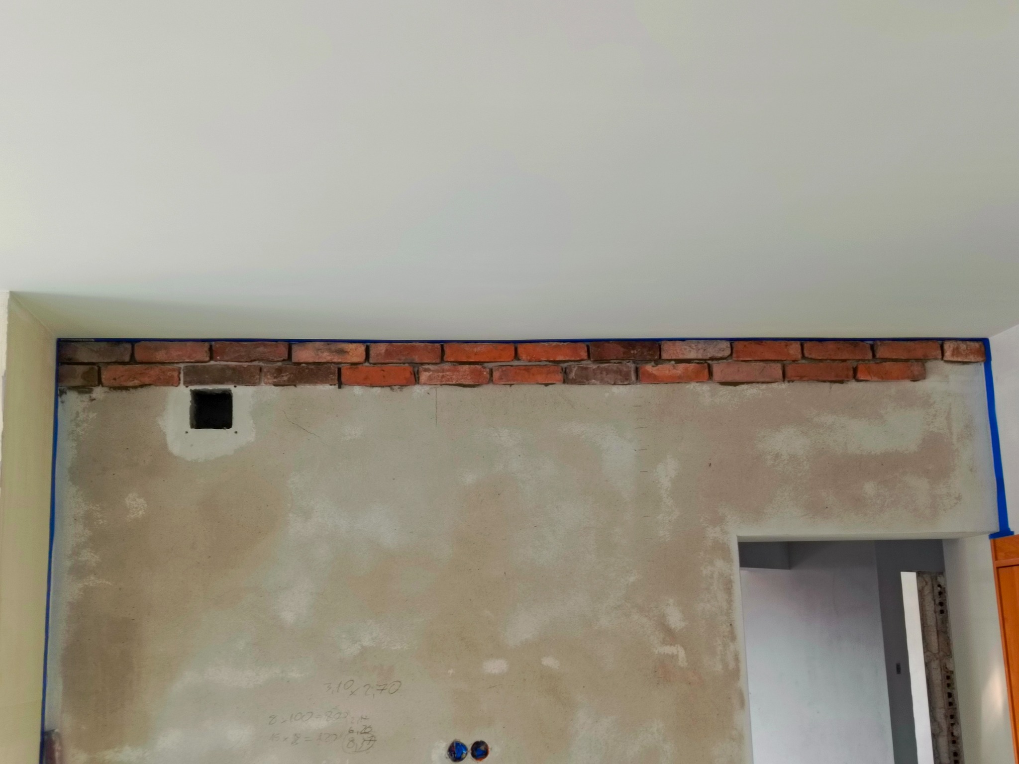 Ściana w trakcie prac wykończeniowych: częściowo położona cegła dekoracyjna obramowana niebieską taśmą malarską, surowy tynk z odręcznymi notatkami i otworem na instalację elektryczną, widoczny...