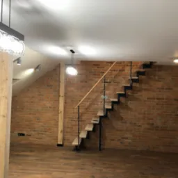 Loftowe wnętrze z drewnianymi schodami na metalowej konstrukcji, ceglaną ścianą i drewnianą kolumną, oświetlone nowoczesnymi lampami.