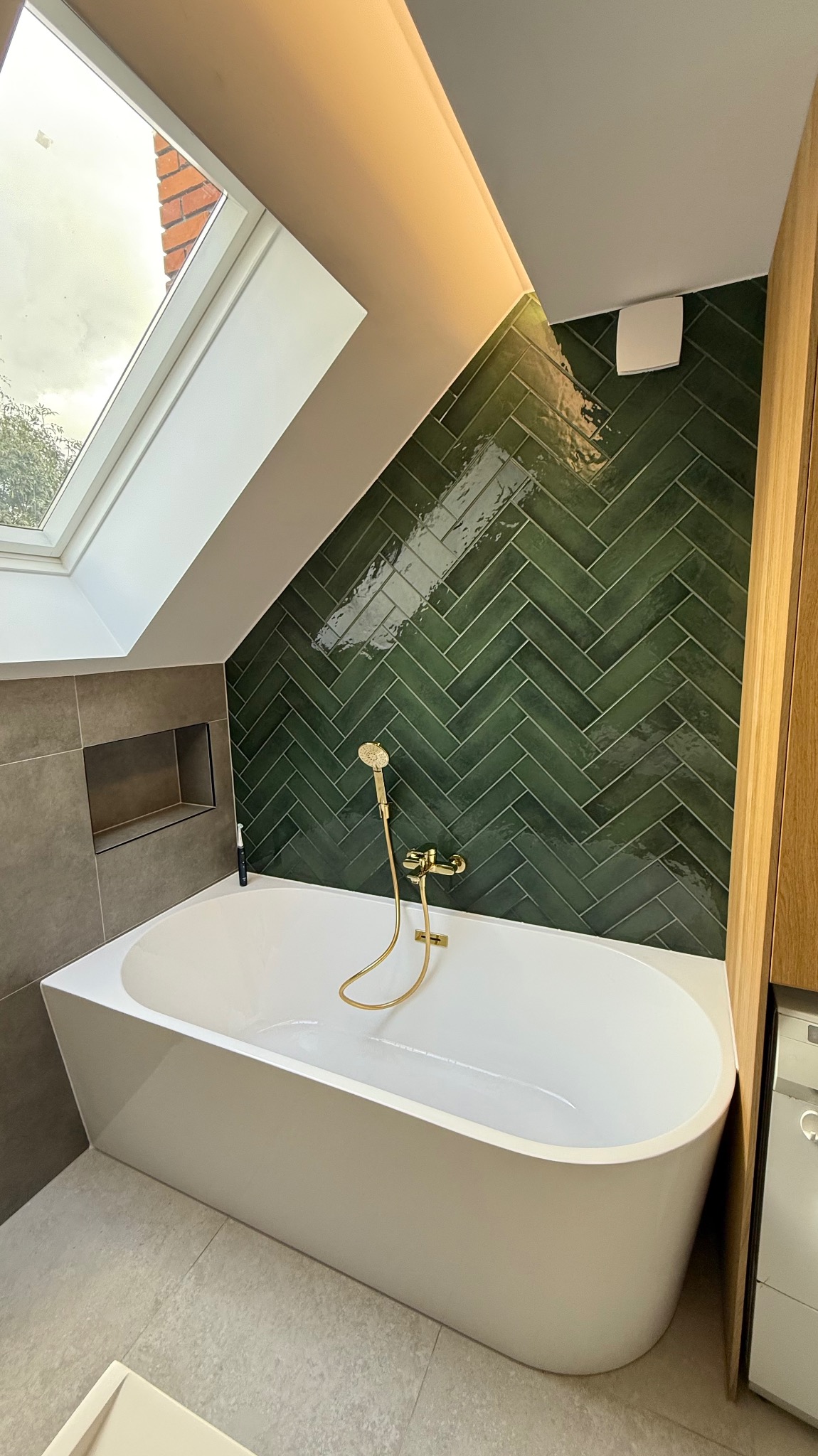 Nowoczesna łazienka na poddaszu z białą wanną, zielonymi płytkami ułożonymi w jodełkę i złotą armaturą. Okno dachowe doświetla wnętrze.