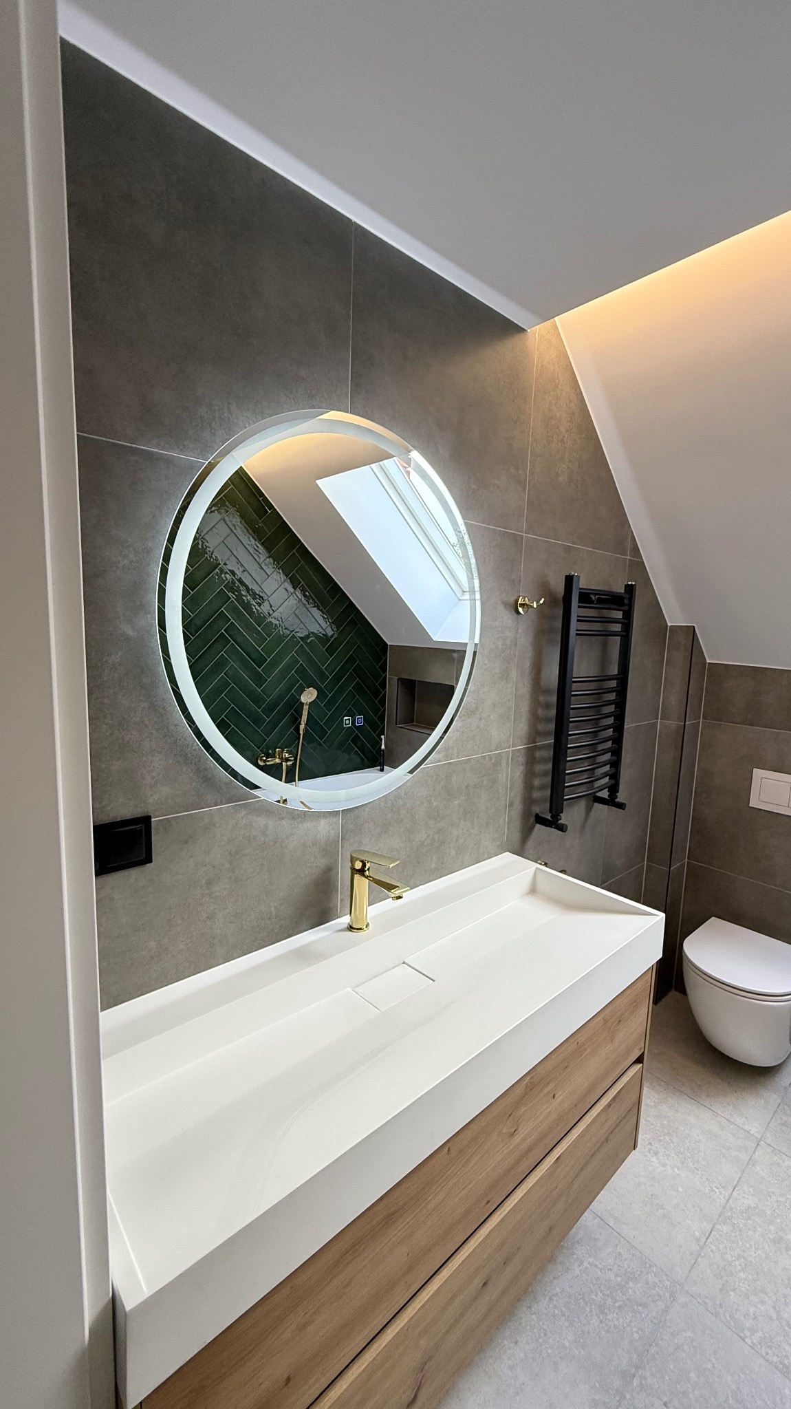 Nowoczesna łazienka na poddaszu z okrągłym lustrem LED odbijającym zieloną mozaikę, białą umywalką z drewnianą szafką i grzejnikiem drabinkowym.