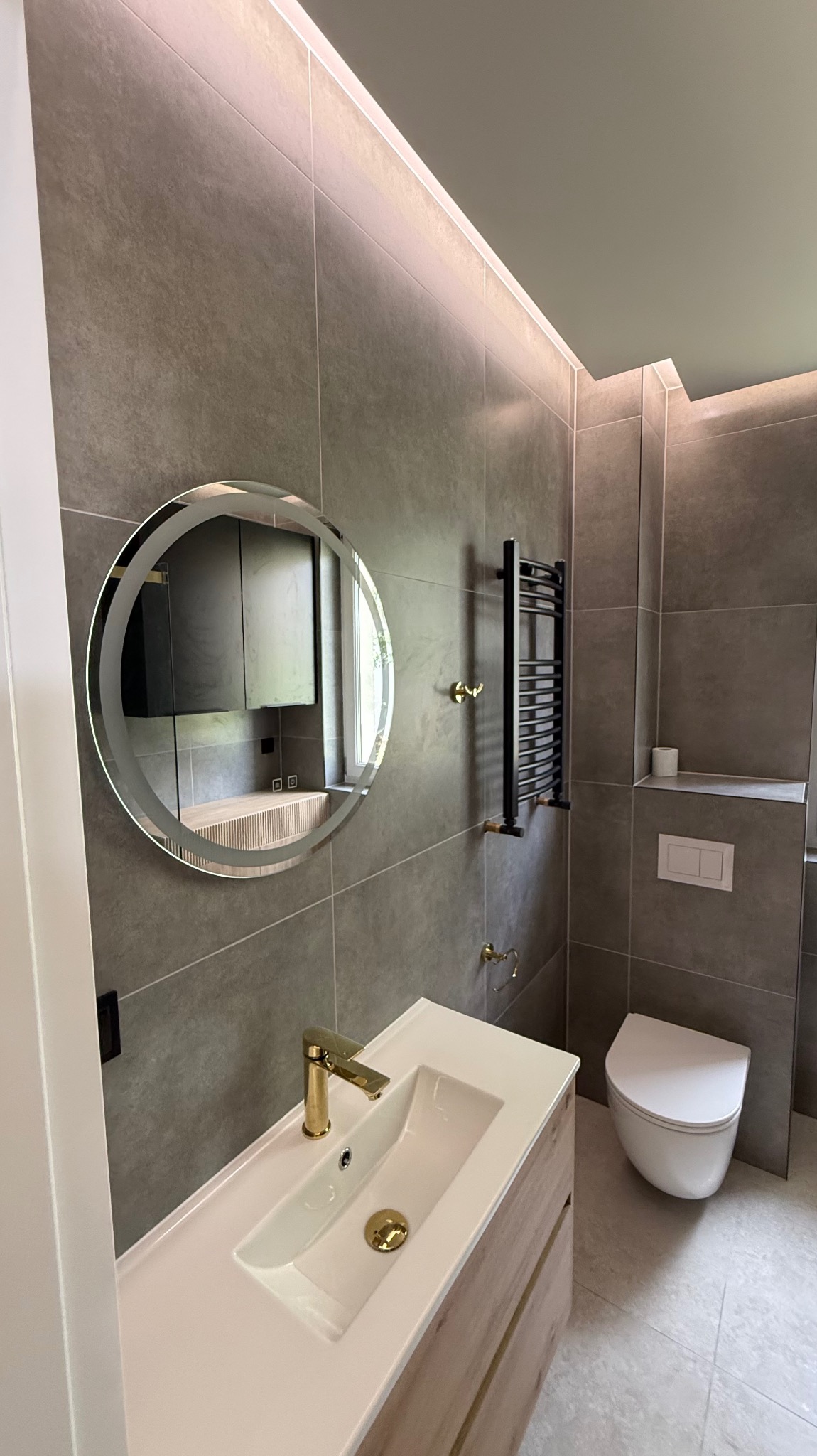 Nowoczesna łazienka z szarymi płytkami, okrągłym lustrem z podświetleniem, złotą armaturą i grzejnikiem drabinkowym. Minimalistyczny design z dbałością o detale.