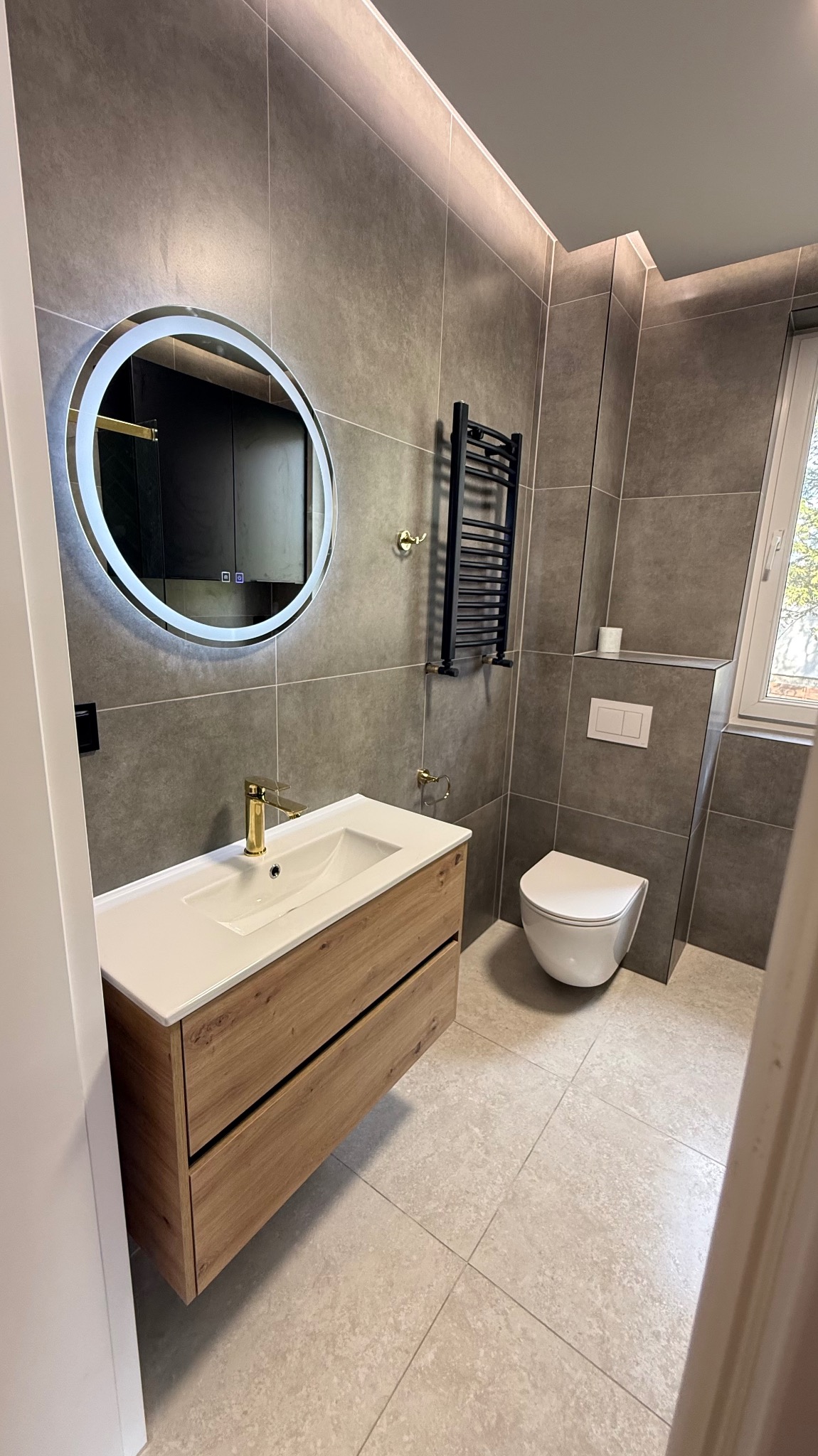 Nowoczesna łazienka z szarymi płytkami, drewnianą szafką pod umywalką z złotą armaturą i okrągłym lustrem z podświetleniem LED. Minimalistyczny design.