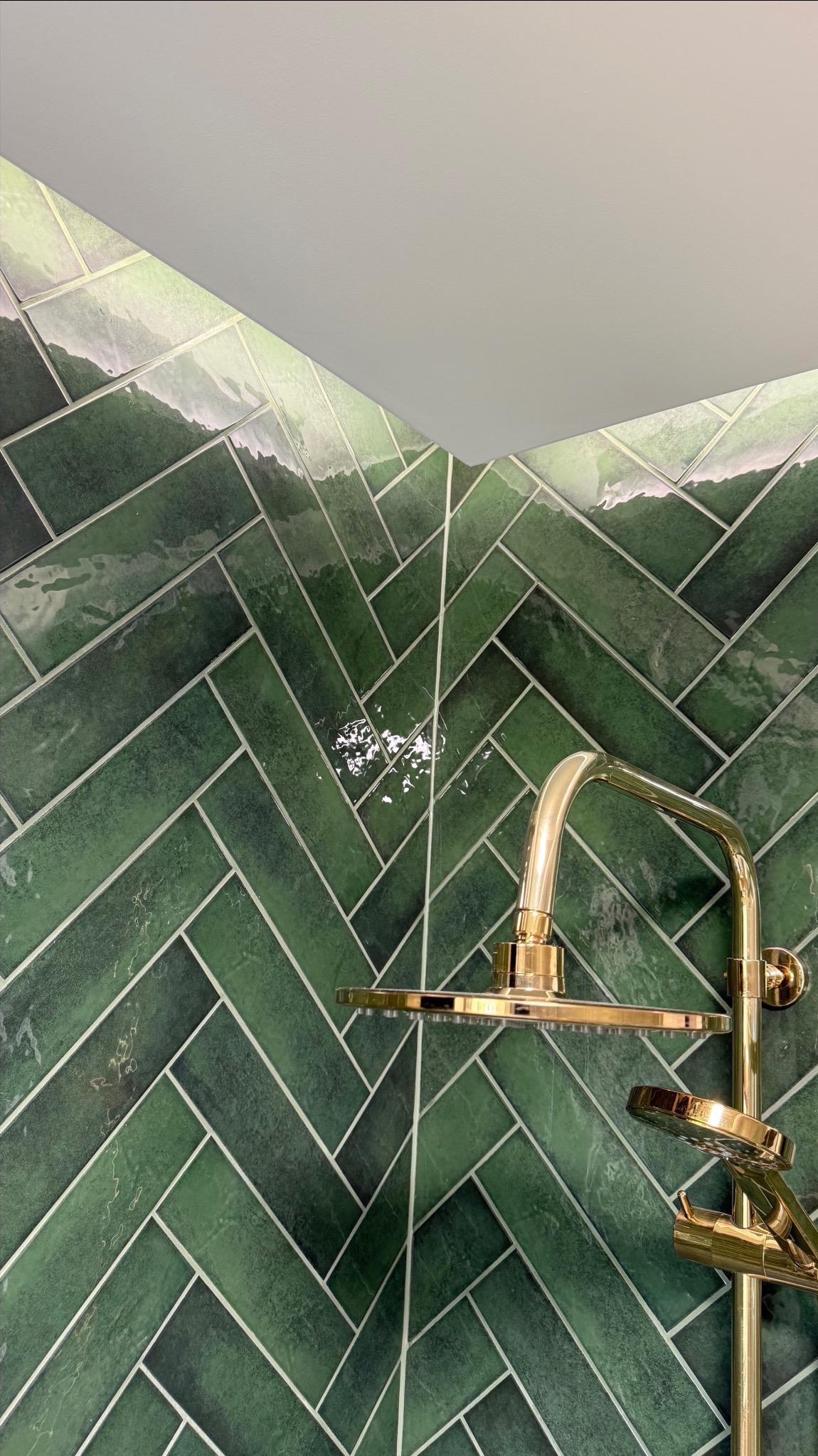 Błyszczące, zielone płytki ułożone w jodełkę na ścianie kabiny prysznicowej z złotą armaturą. Nowoczesny design łazienki z dbałością o detale.