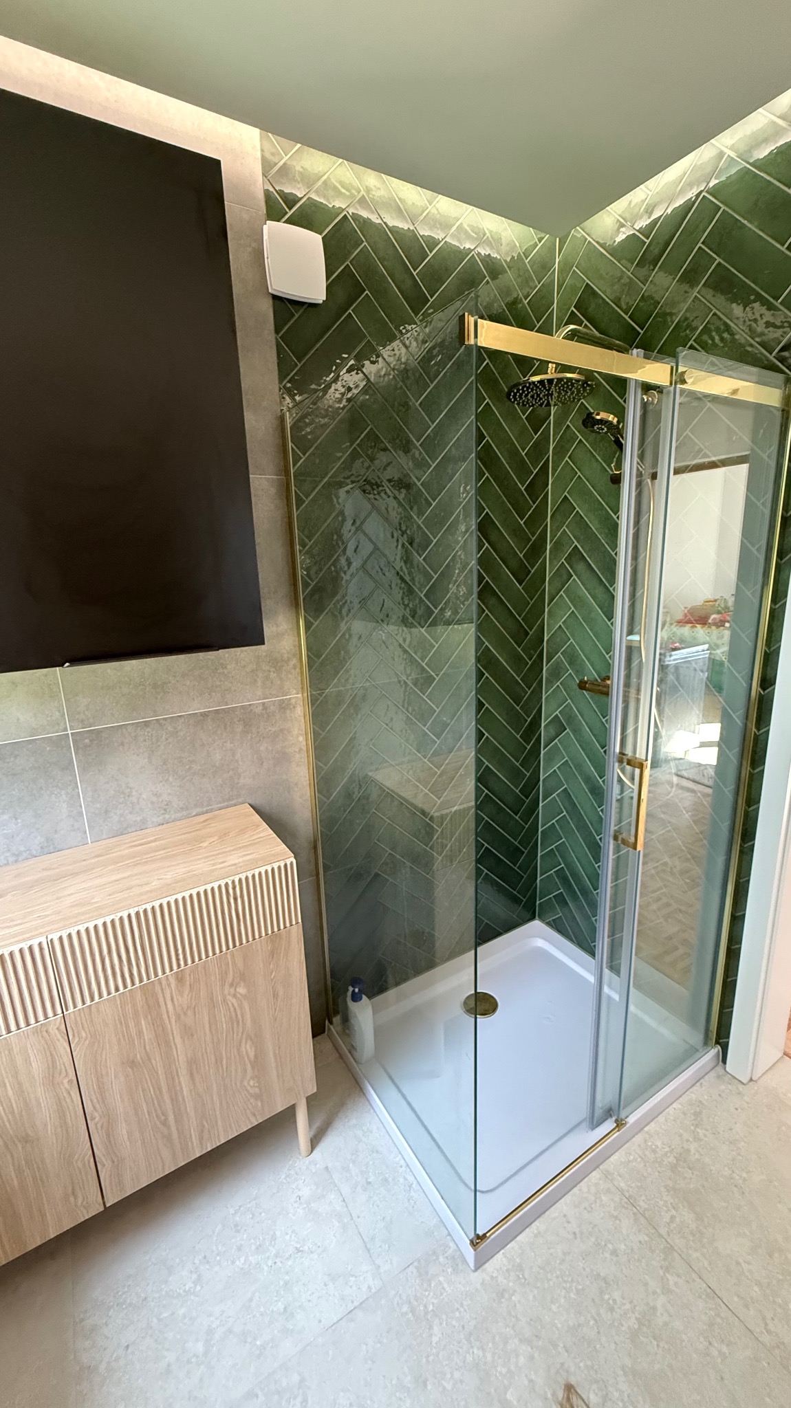 Nowoczesna łazienka z kabiną prysznicową w złotej ramie i zielonymi płytkami ułożonymi w jodełkę. Jasna szafka pod umywalką i szare płytki na ścianie.