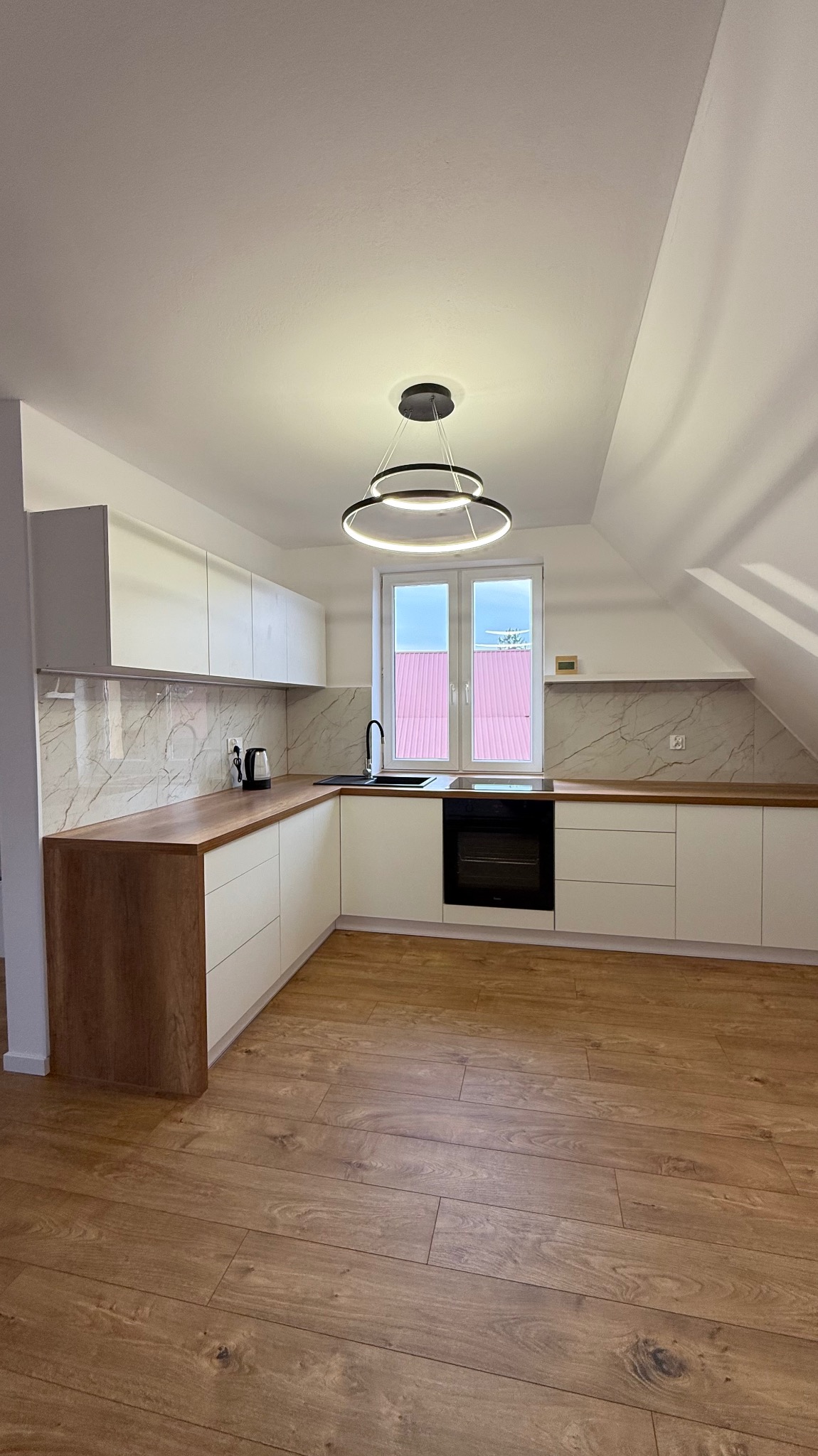 Nowoczesna kuchnia na poddaszu z białymi szafkami, drewnianym blatem i panelami podłogowymi. Okno z widokiem na czerwony dach. Minimalistyczna lampa sufitowa.