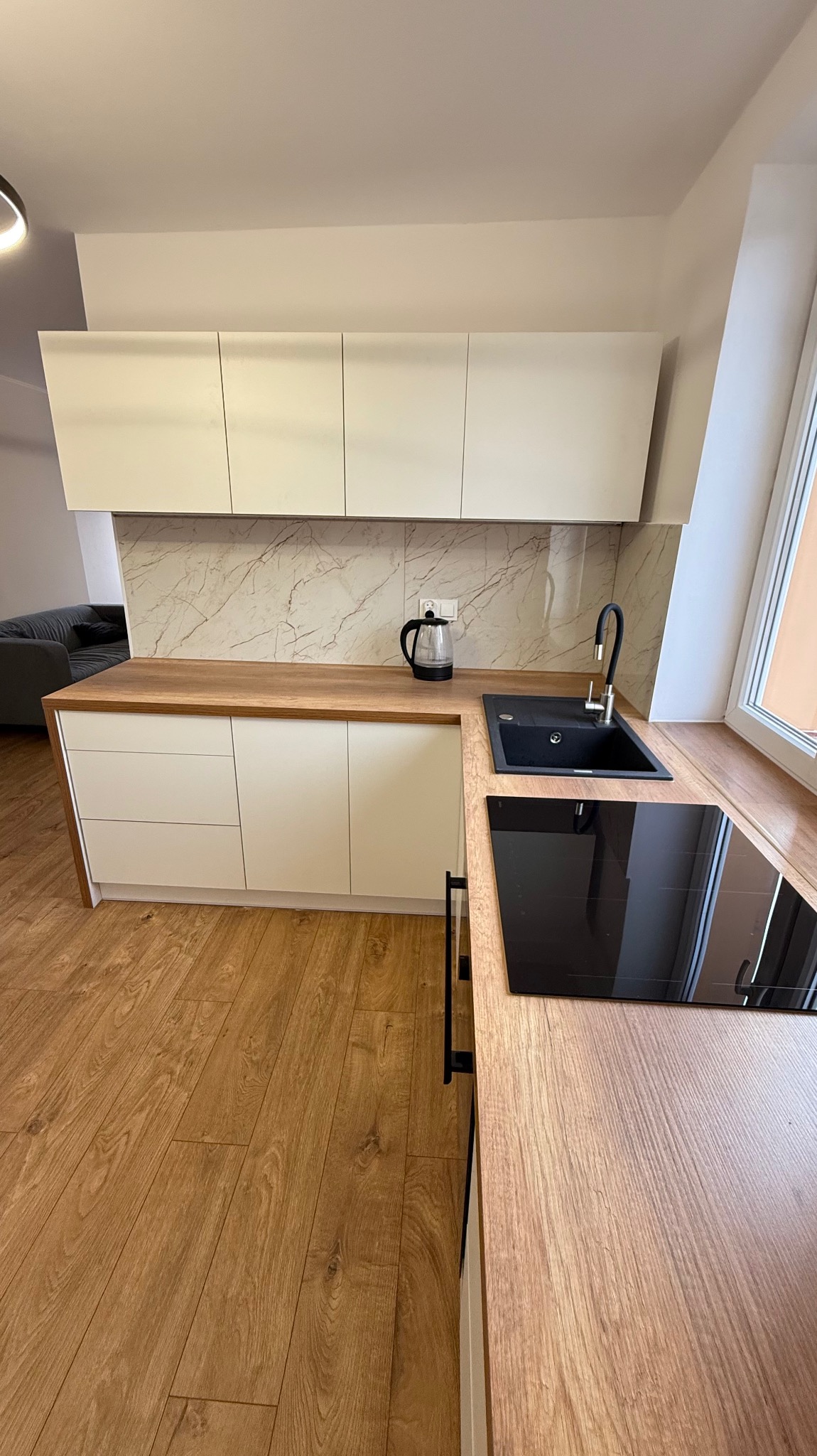 Nowoczesna kuchnia z białymi szafkami, drewnianym blatem i marmurową ścianą. Czarny zlew i płyta indukcyjna. Jasne panele podłogowe. Minimalistyczny design.