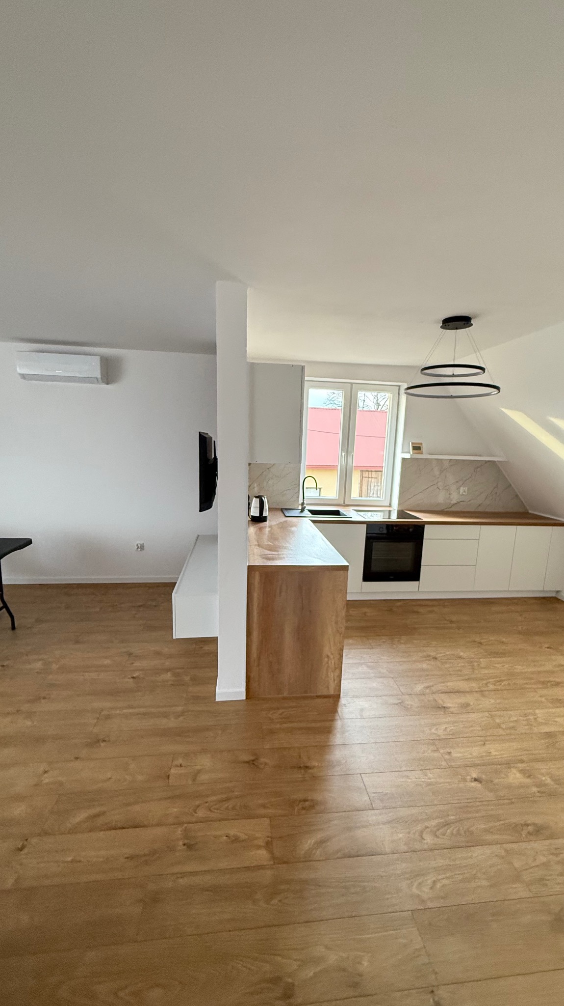 Jasne, przestronne wnętrze po remoncie: drewniana podłoga, białe meble kuchenne z marmurowym blatem, nowoczesne oświetlenie. Widok na okno i fragment ściany z telewizorem.