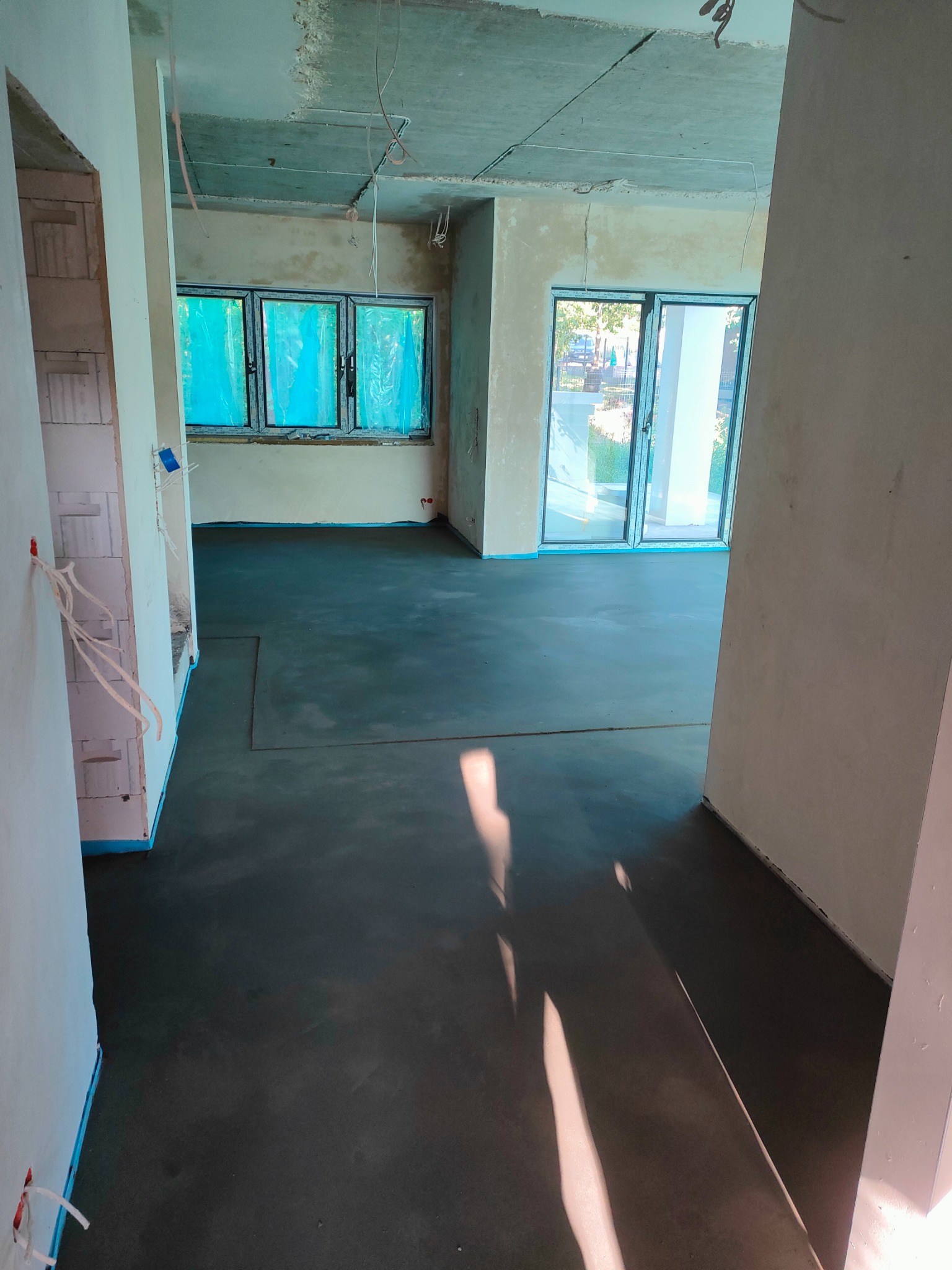 Wnętrze w stanie deweloperskim z wylaną, świeżą posadzką, zabezpieczone okna folią, widoczne kable wystające ze ściany.