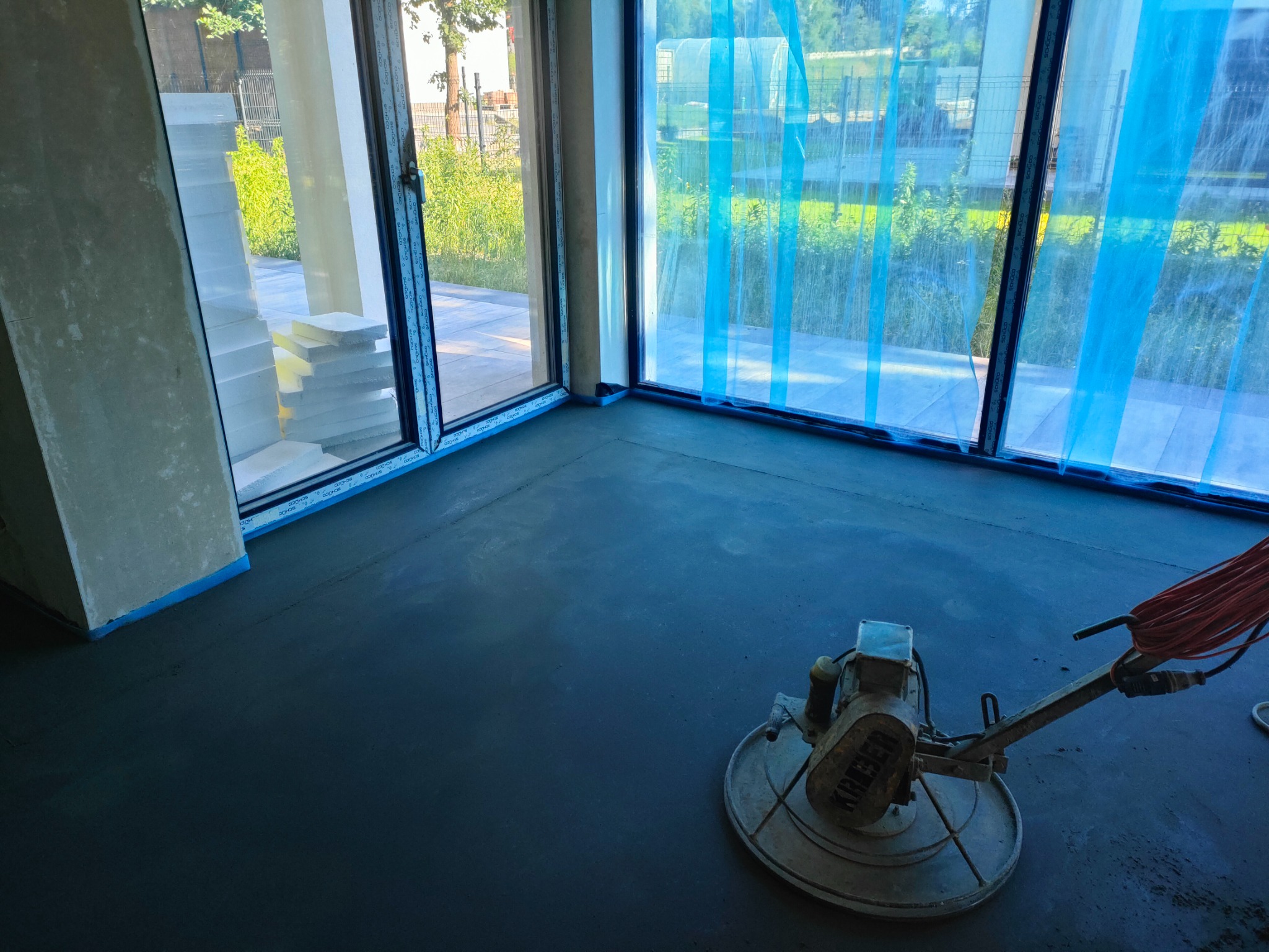 Szlifowanie jastrychu maszyną Kreber w jasnym wnętrzu z dużymi oknami zabezpieczonymi folią, widoczne przygotowane płyty izolacyjne przy ścianie.