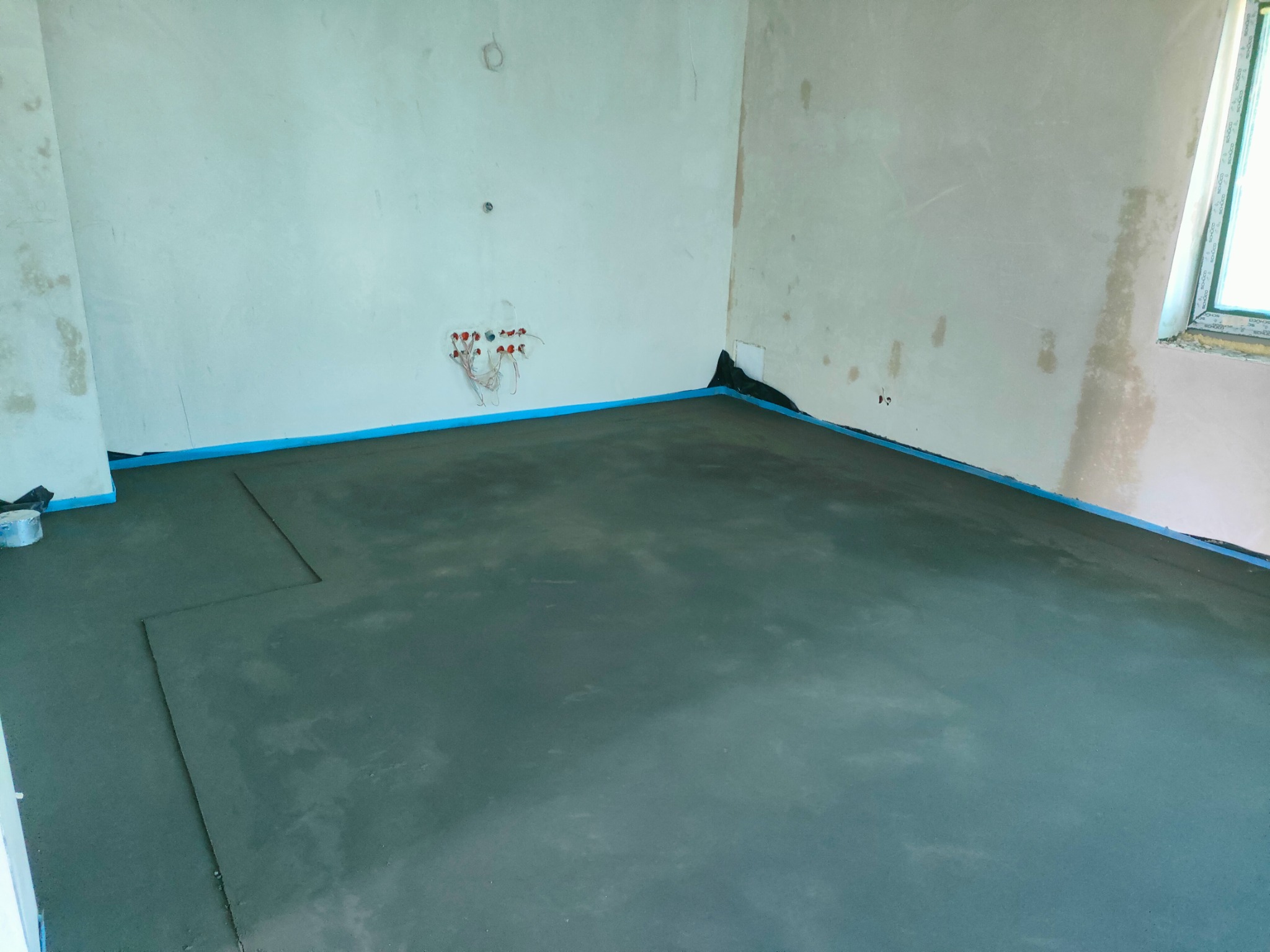 Świeżo wylana, mokra posadzka w pomieszczeniu, zabezpieczona niebieską taśmą przy ścianach i czarną folią w narożnikach. Widoczne wystające kable elektryczne.