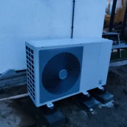 Biała zewnętrzna jednostka klimatyzacji zamontowana na betonowych blokach przy ścianie budynku z widocznymi kablami elektrycznymi.