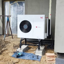 Marek kalkowski Instalator - Biała pompa ciepła Saunier Duval zamontowana na czarnej konstrukcji wsporczej, z widocznymi przewodami i narzędziami na niebieskiej szmatce, na tle surowej elewacji budynku w trakcie wykończenia.