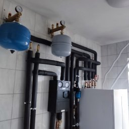 Marek kalkowski Instalator - Kompleksowa instalacja grzewcza w pomieszczeniu z białymi kafelkami, widoczne czarne rury, zbiornik buforowy, manometry i inne elementy instalacji.