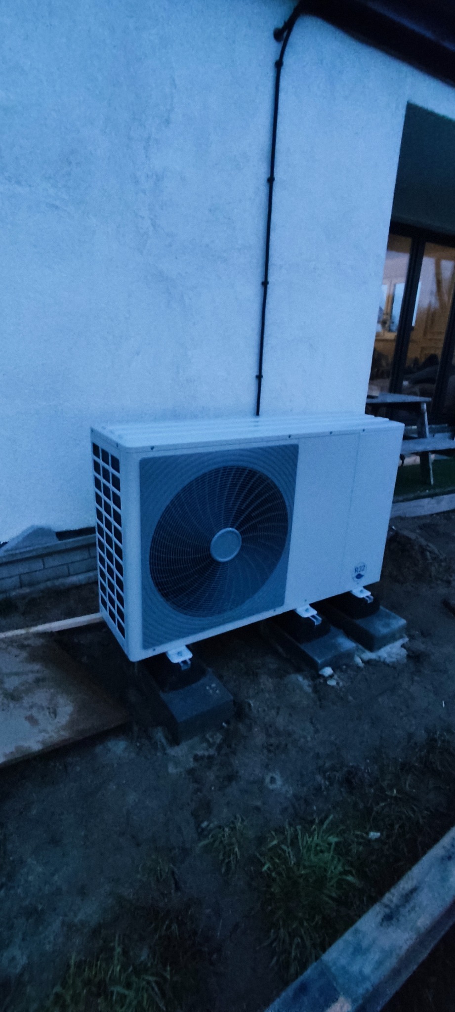 Biała zewnętrzna jednostka klimatyzacji zamontowana na betonowych blokach przy ścianie budynku z widocznymi kablami elektrycznymi.