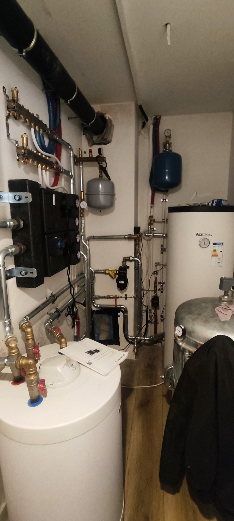 Złożona instalacja grzewcza w kotłowni, widoczne liczne rury, zbiorniki wyrównawcze, rozdzielacze, filtry, zawory oraz dokumentacja techniczna leżąca na białym zasobniku. Czarna kurtka w prawym...