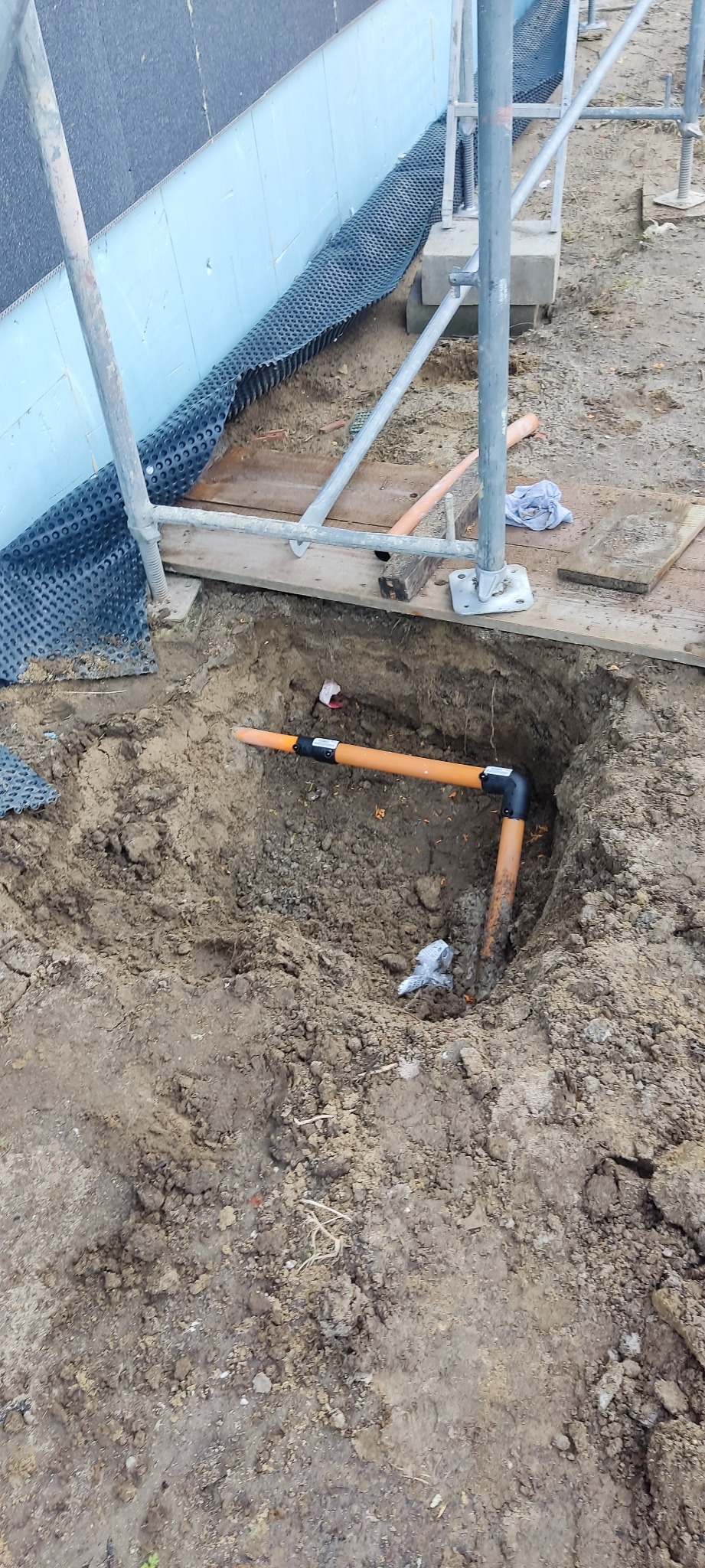 Wykop z ułożonymi rurami kanalizacyjnymi z kolankami, widoczne rusztowanie i izolacja fundamentów budynku w tle.