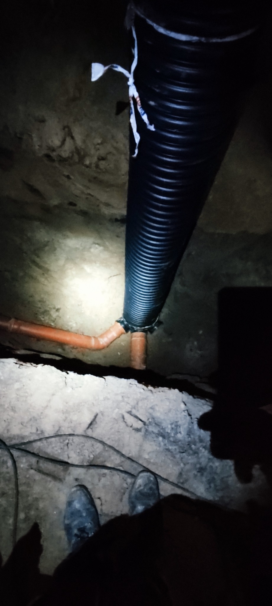 Czarna karbowana rura odpływowa połączona z pomarańczową rurą kanalizacyjną w wykopie, widoczne buty robotnika i oświetlenie punktowe.