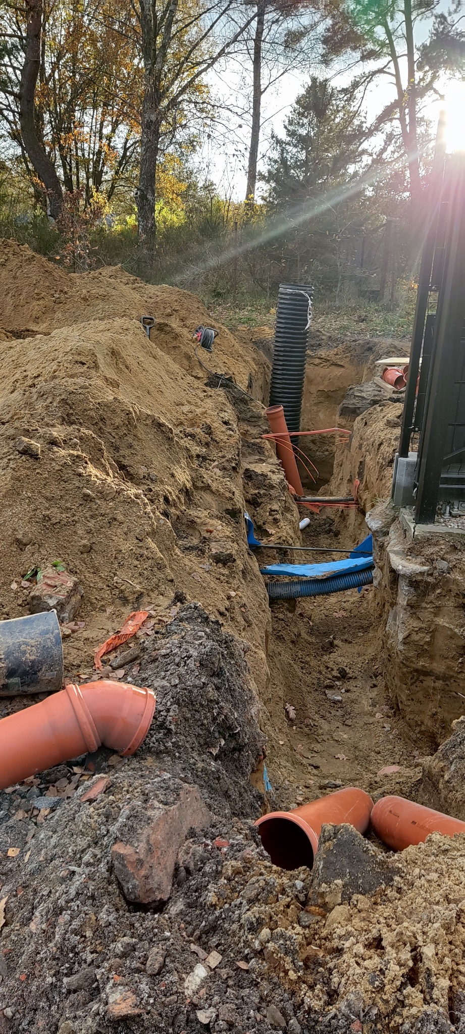Wykop z ułożonymi rurami kanalizacyjnymi z tworzywa sztucznego w ziemi, widoczne elementy przyłącza wodno-kanalizacyjnego oraz rury drenarskie, ziemia częściowo zasypana, w tle drzewa i krzewy.