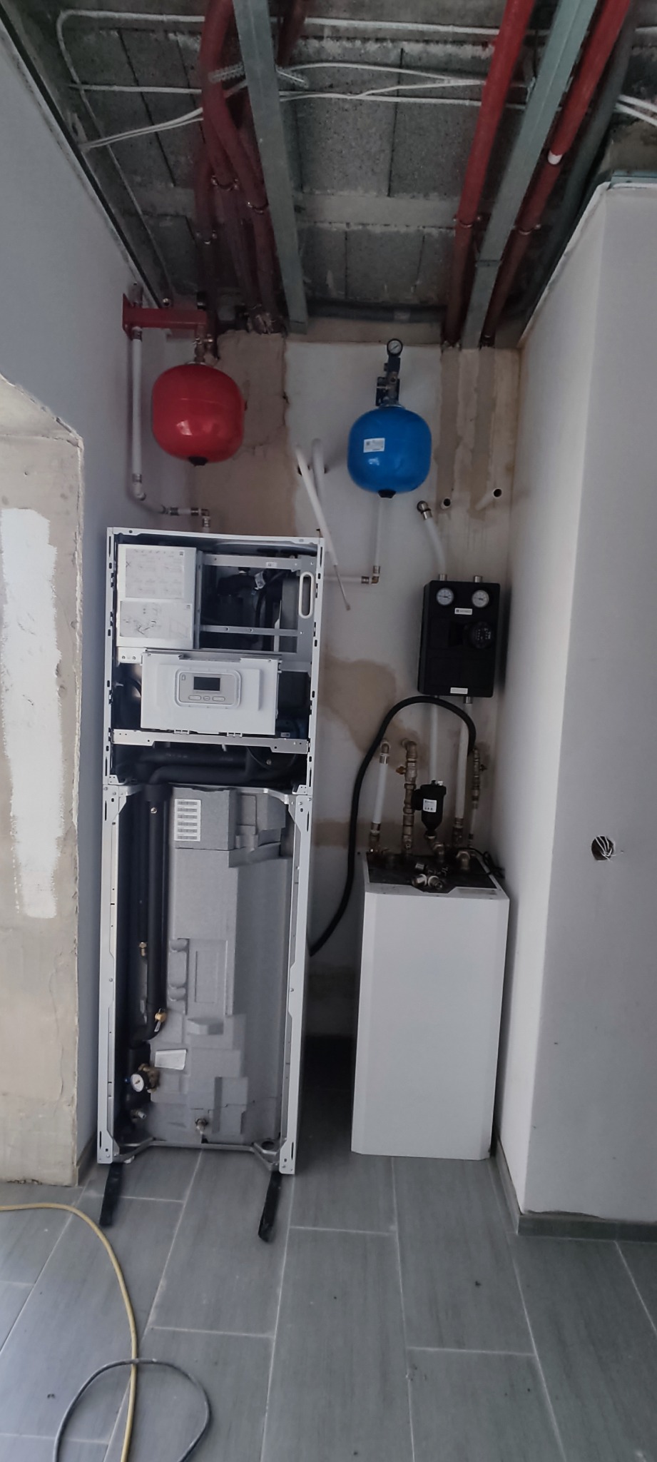 Wnętrze w trakcie remontu z widoczną instalacją centralnego ogrzewania: piec dwufunkcyjny w trakcie montażu, czerwony i niebieski zbiornik wyrównawczy, rury miedziane pod sufitem, surowy tynk...