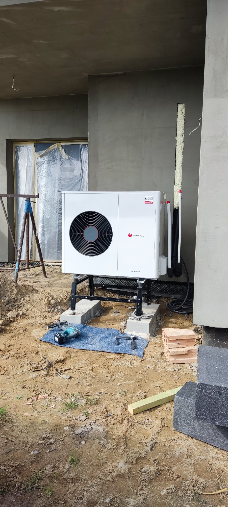 Biała pompa ciepła Saunier Duval zamontowana na czarnej konstrukcji wsporczej, z widocznymi przewodami i narzędziami na niebieskiej szmatce, na tle surowej elewacji budynku w trakcie wykończenia.