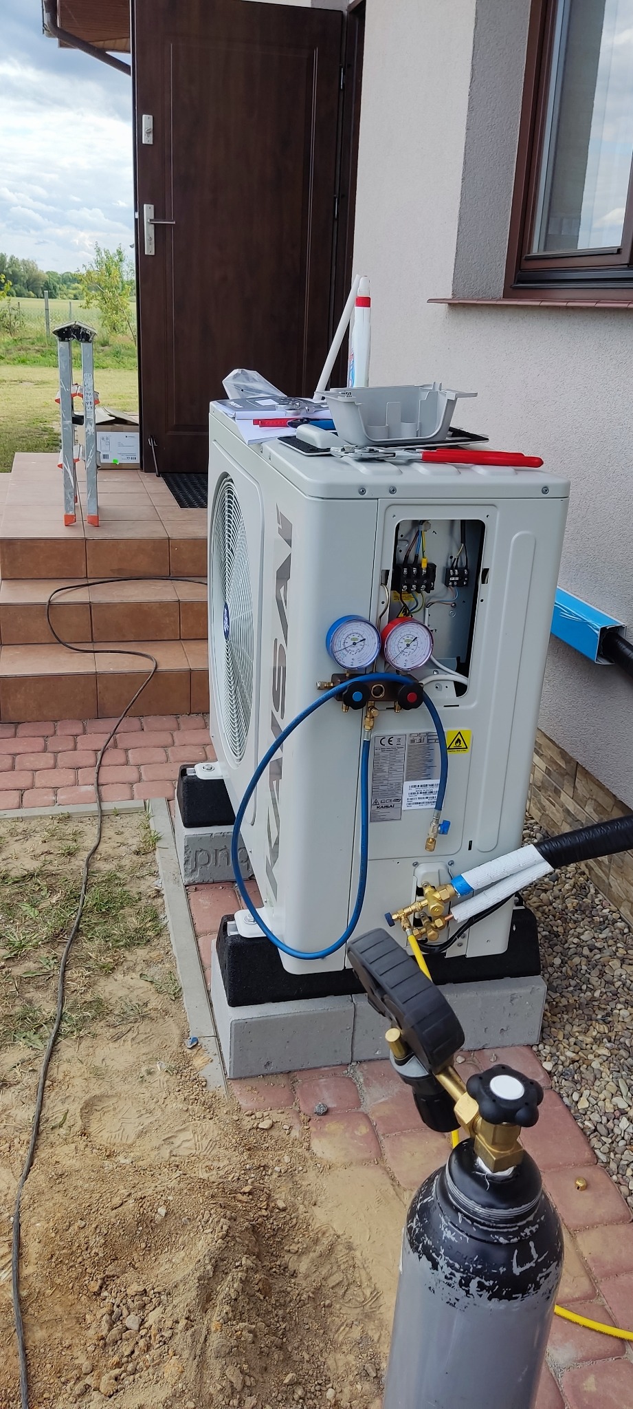 Biała pompa ciepła marki Kaisai, zamontowana na betonowych bloczkach przed domem, podłączona do butli z gazem za pomocą manometrów i węży, z otwartą obudową ukazującą wewnętrzne okablowanie.