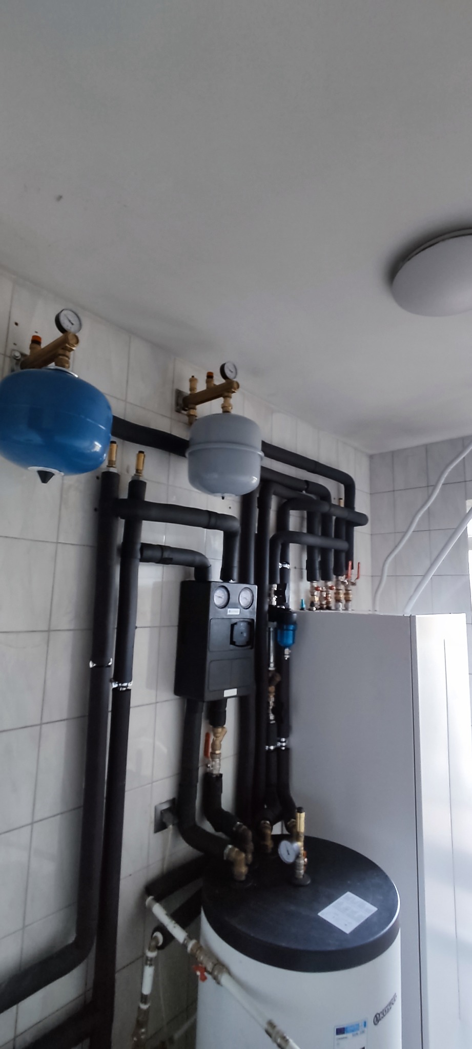 Kompleksowa instalacja grzewcza w pomieszczeniu z białymi kafelkami, widoczne czarne rury, zbiornik buforowy, manometry i inne elementy instalacji.