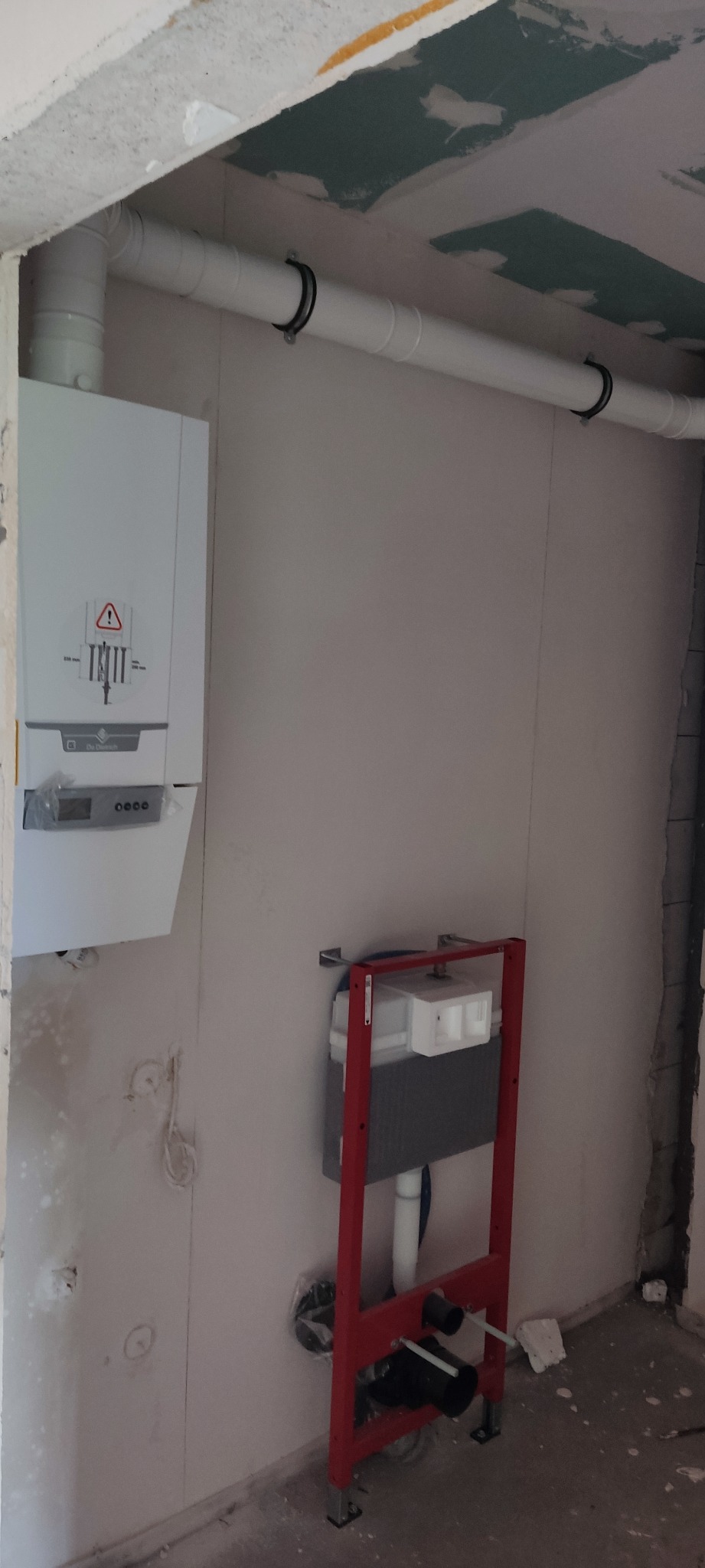 Biały piec gazowy zamontowany na ścianie w surowym stanie, z widocznymi rurami i instalacją podtynkową WC ze stelażem w kolorze czerwonym, w trakcie prac wykończeniowych.